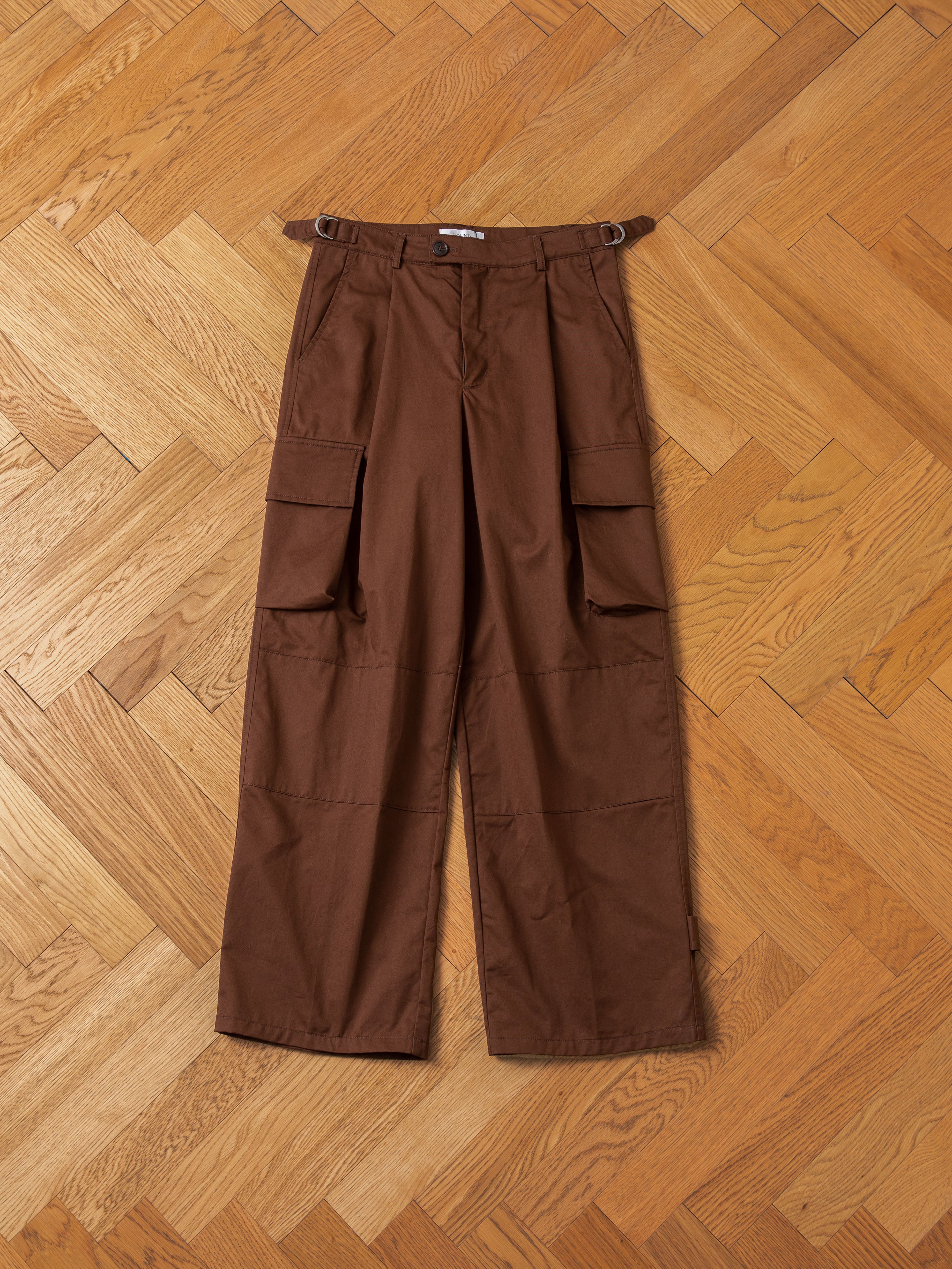 A pair of FOUND Pleated Pocket Trousers with side cargo pockets laid flat on a wooden herringbone floor.