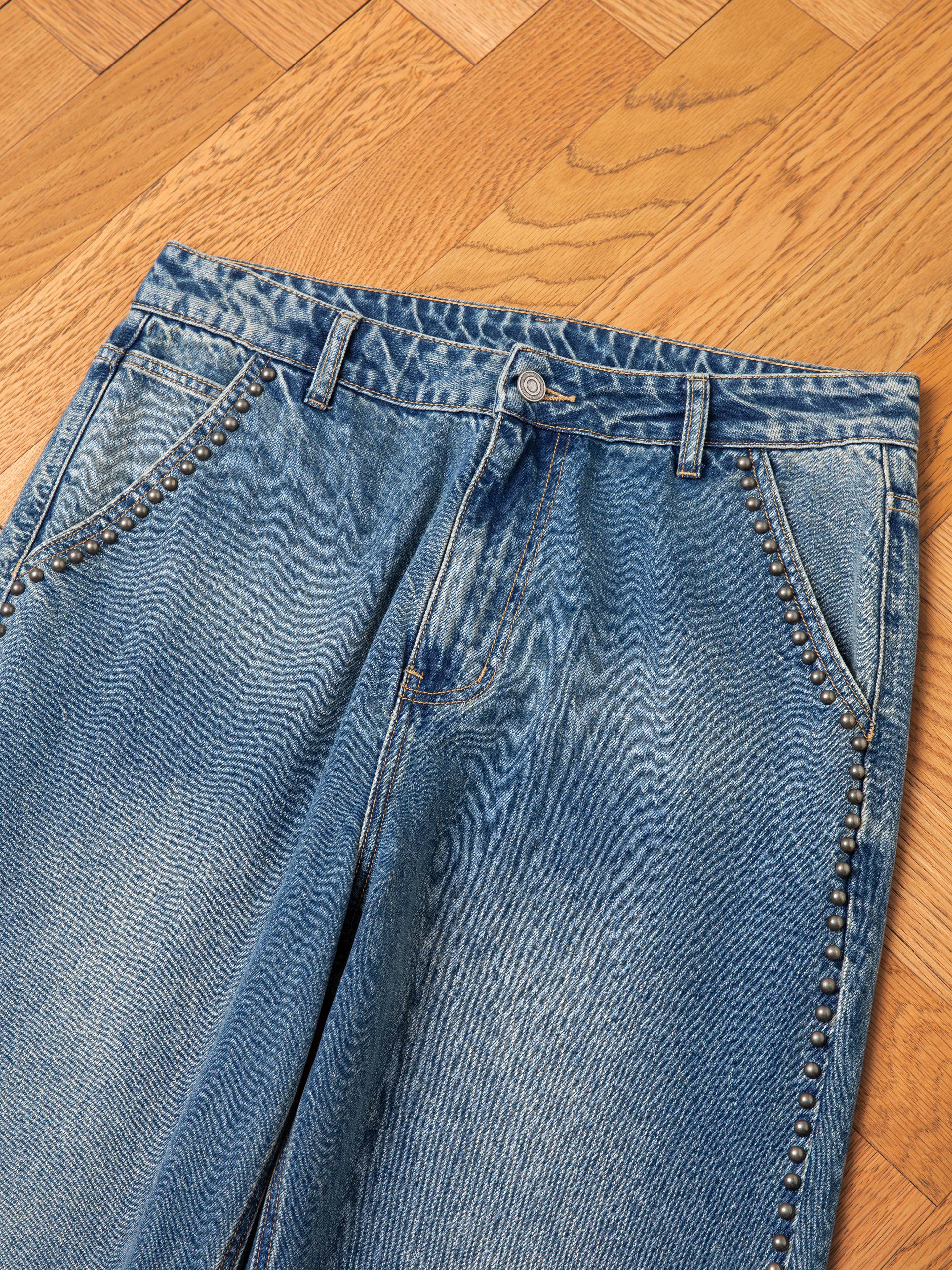 A pair of FOUND Azar Studded Jeans in a vintage blue wash, featuring antique matte silver studs on the front pockets and along the side seams, laid flat on a wooden parquet floor.