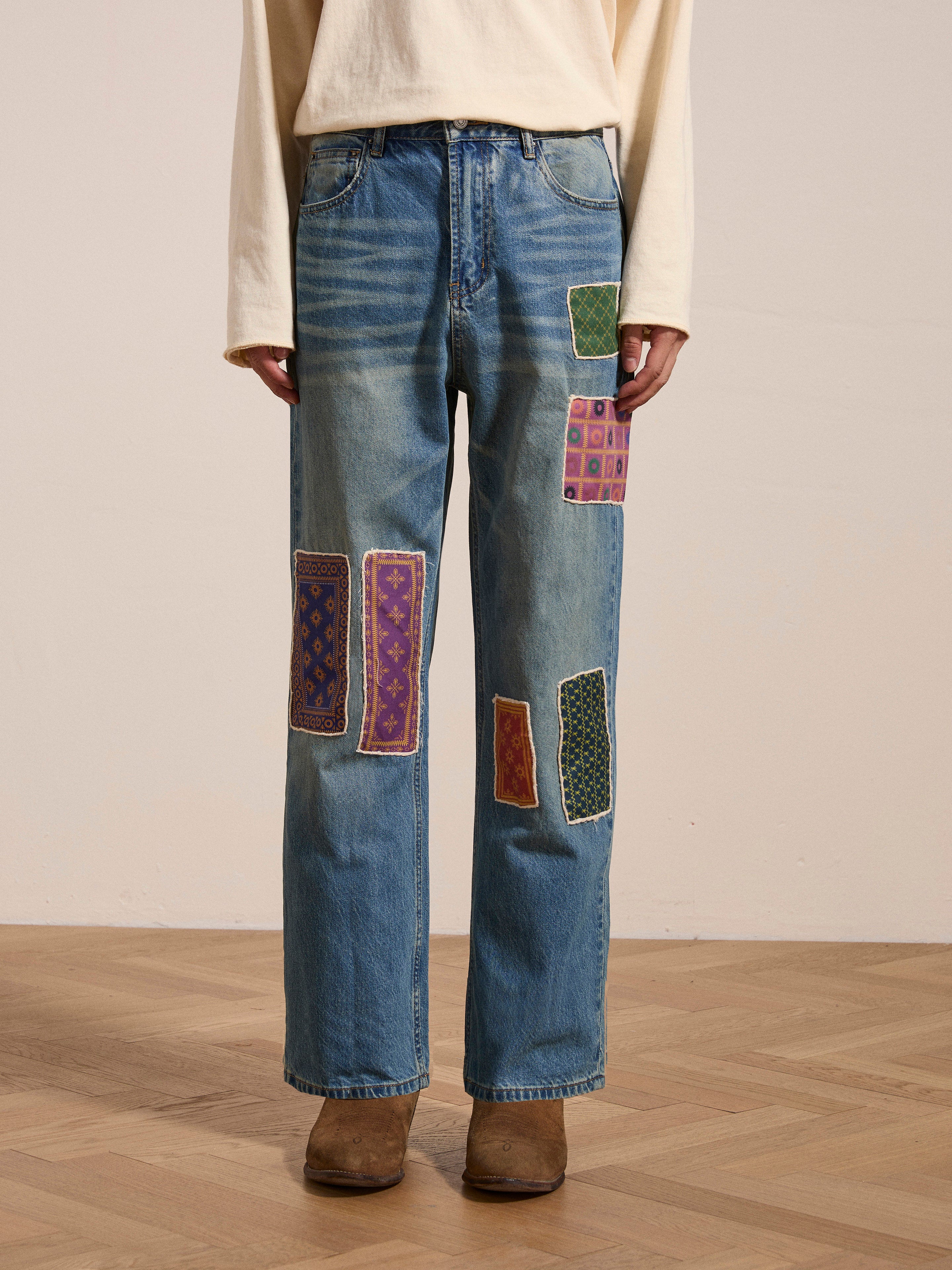 A person wearing a light-colored long-sleeve shirt and Found's Kantha Patchwork Jeans, adorned with colorful South Asian inspired patches, stands on a wooden floor. Only their torso and legs are visible, and they are wearing brown shoes.