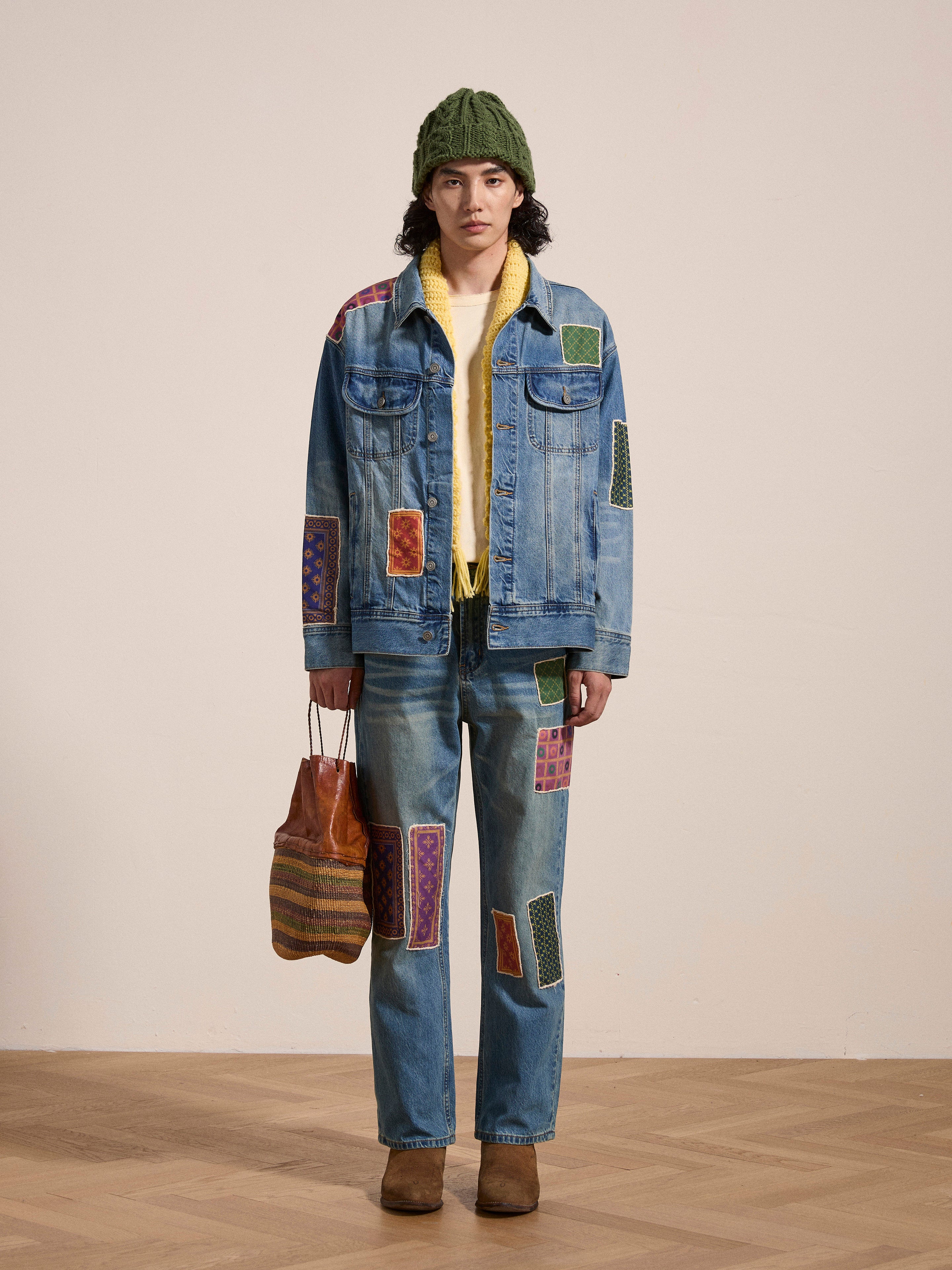 A person stands indoors wearing a FOUND Kantha Patchwork Denim Trucker Jacket with colorful patterns, a green beanie, a yellow inner shirt, holding a brown and striped handbag, and brown shoes.