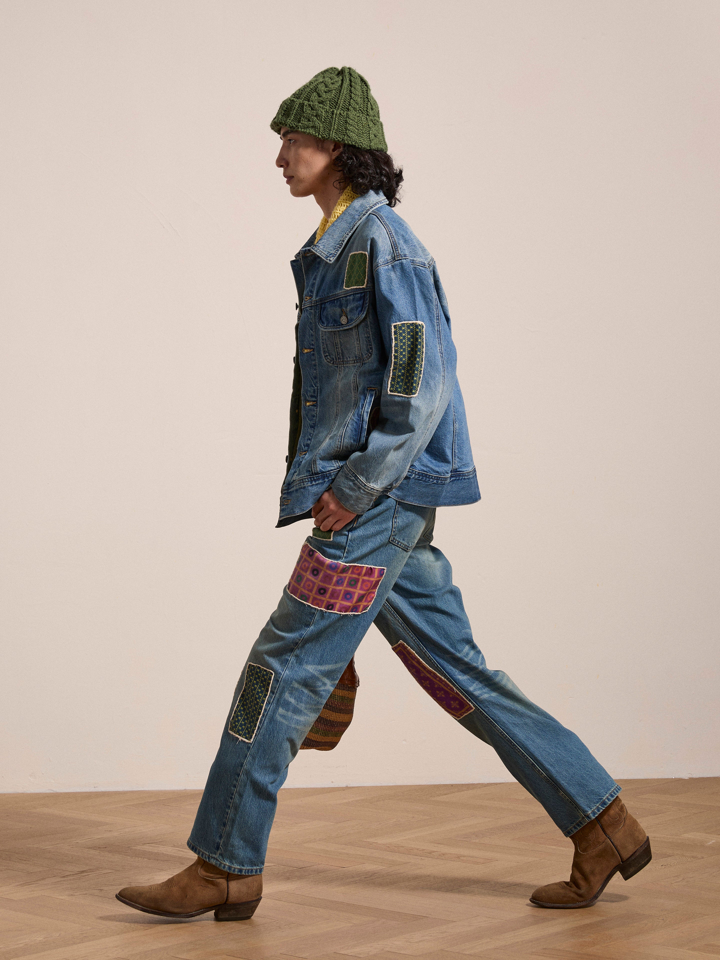 A person walks indoors wearing a green knit hat, a blue patchwork denim jacket with South Asian inspired patches, Found's Kantha Patchwork Jeans in a vintage blue wash, and brown boots.