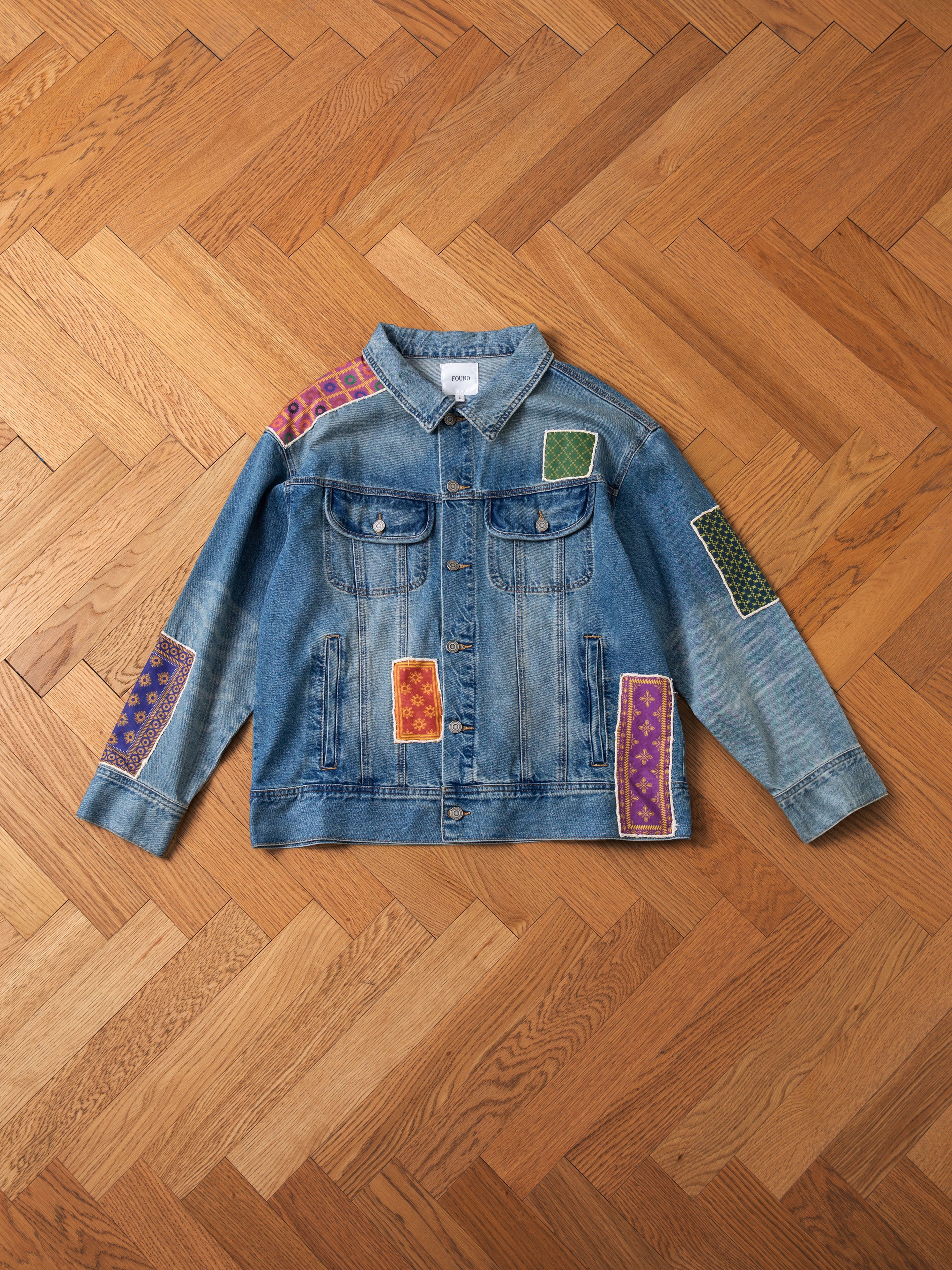 The FOUND Kantha Patchwork Denim Trucker Jacket, adorned with vibrant South Asian inspired patches, is laid flat on a wooden parquet floor. It features button closures and two chest pockets.