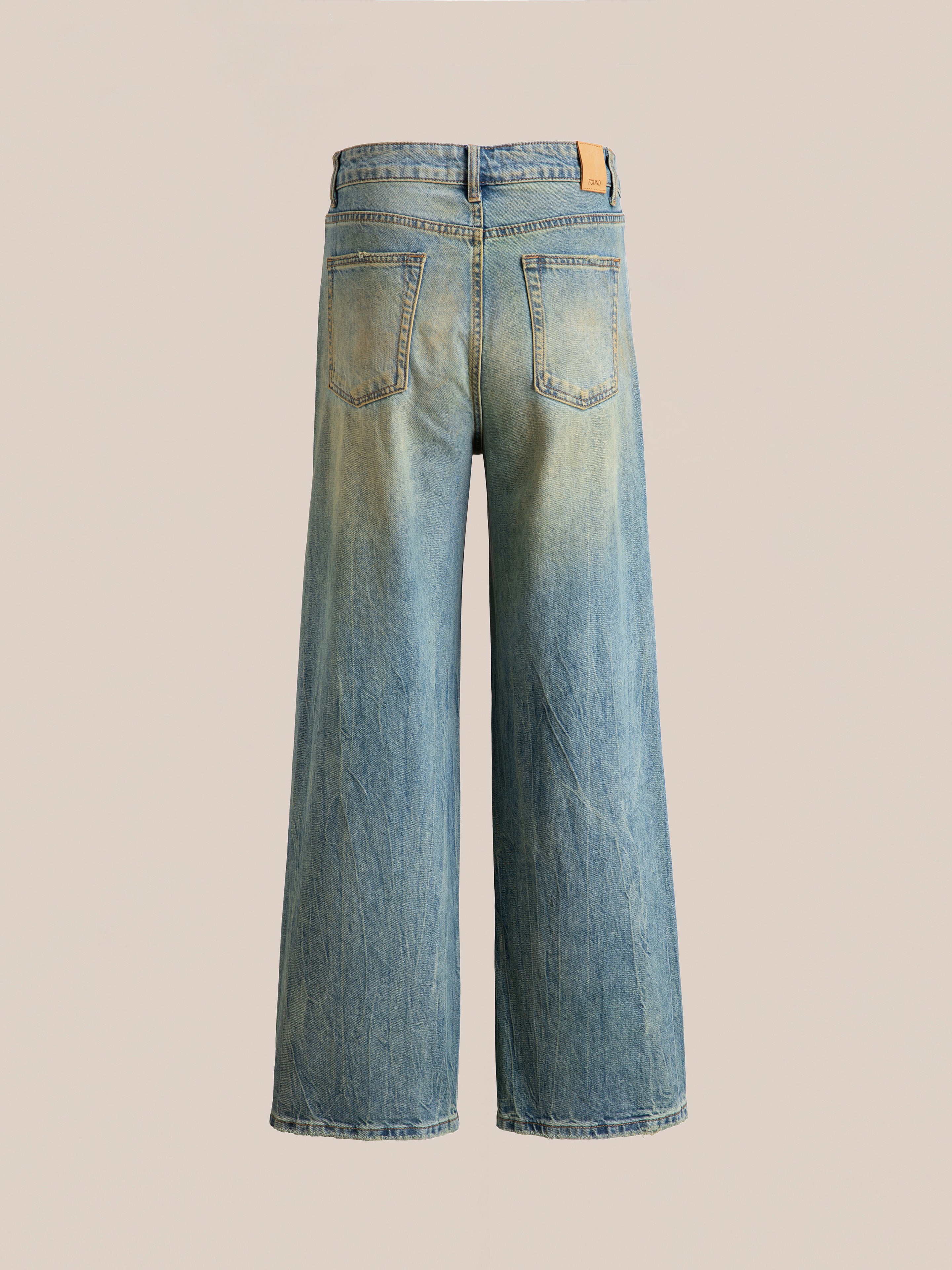 Back view of the Lacy Baggy Jeans by FOUND, showcasing a pair of wide-leg blue jeans in a dirty blue wash with light distressing, two back pockets, and a leather patch on the waistband, set against a plain beige background.