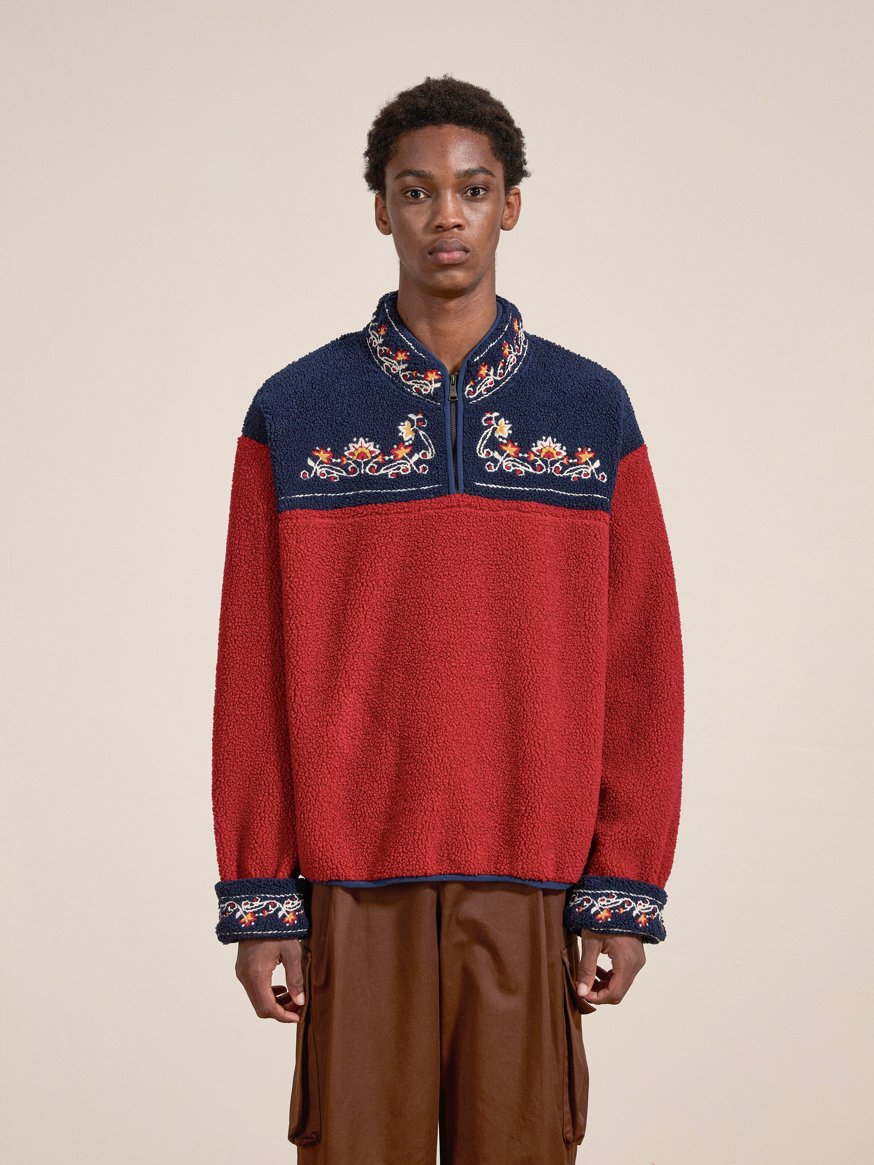 A person stands wearing the FOUND Pahari Floral Embroidered Sherpa Pullover with brown pants, looking at the camera against a plain background. The pullover features subtle Pahari style embroidery that adds an intricate touch to their outfit.