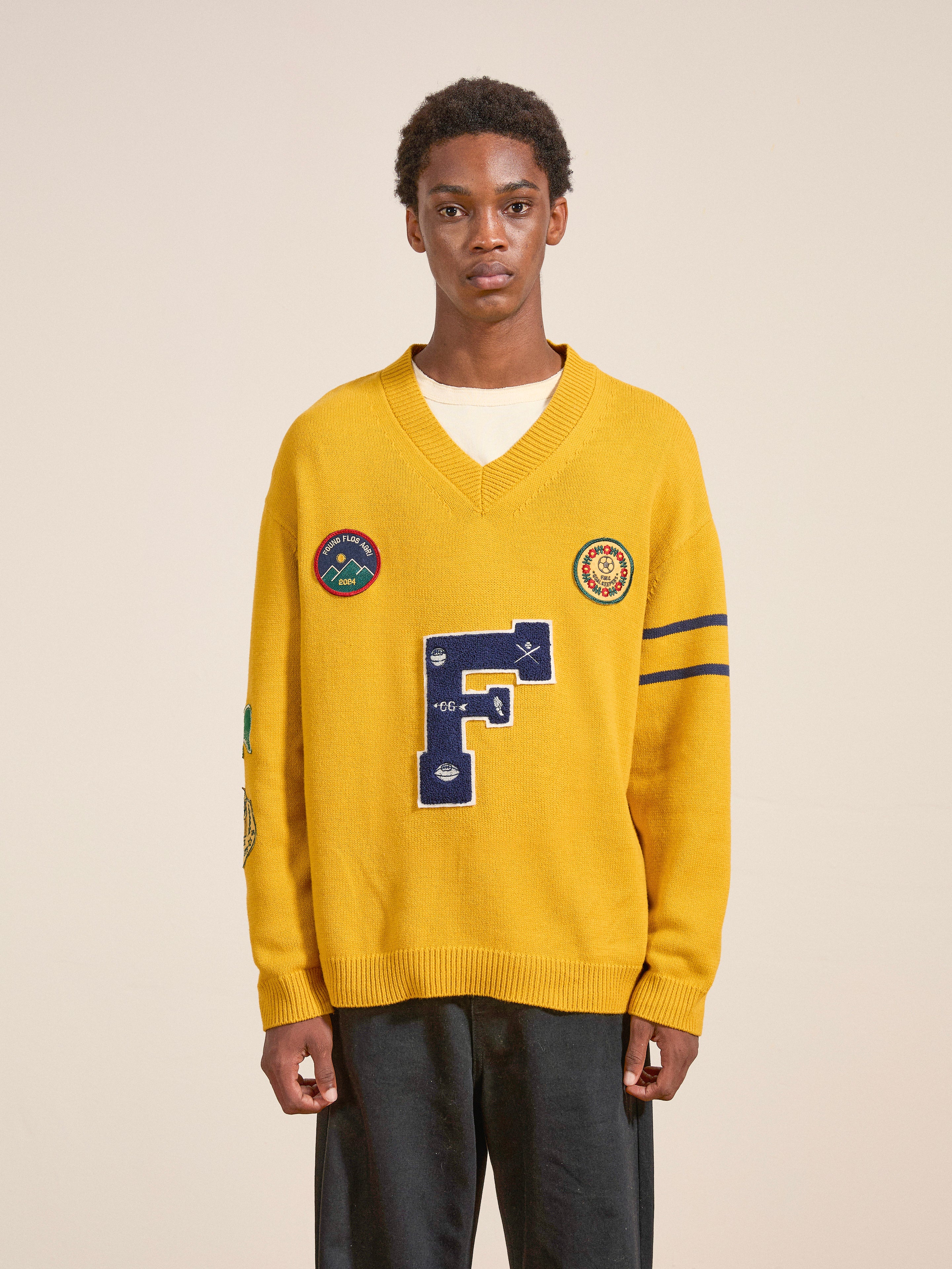 A person stands against a plain background, wearing a yellow Arrow-Wood Varsity Knit Sweater by FOUND, featuring patches that include a large blue "F" and other decorative symbols, paired with dark pants for that vintage look.