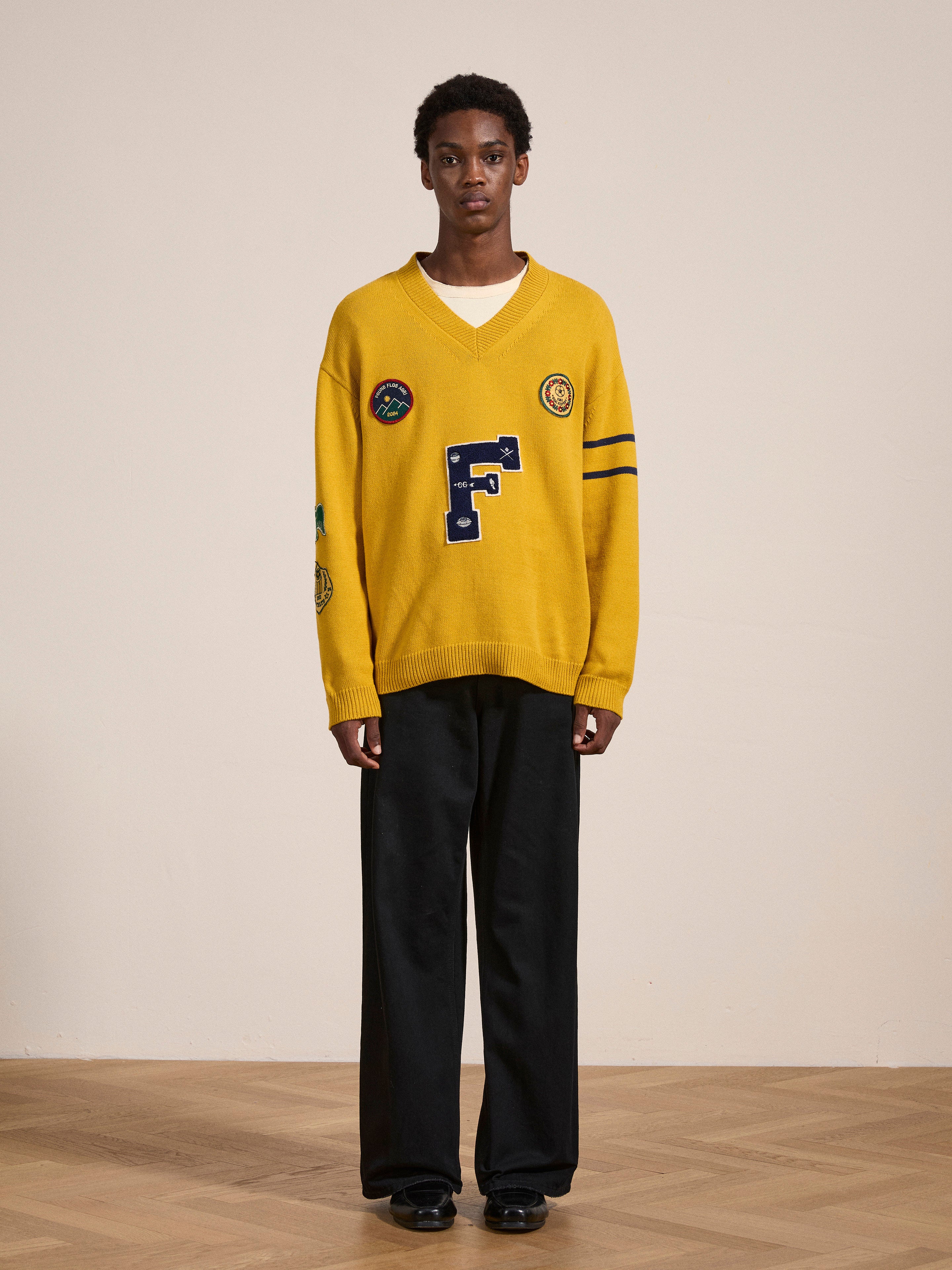 A person stands indoors wearing an Arrow-Wood Varsity Knit Sweater with patches and a large "F" on the front, over a white shirt and black wide-leg pants. The wooden floor complements the beige background wall, creating an Ivy League wardrobe feel. The sweater, crafted by FOUND, adds a vintage touch to the ensemble.