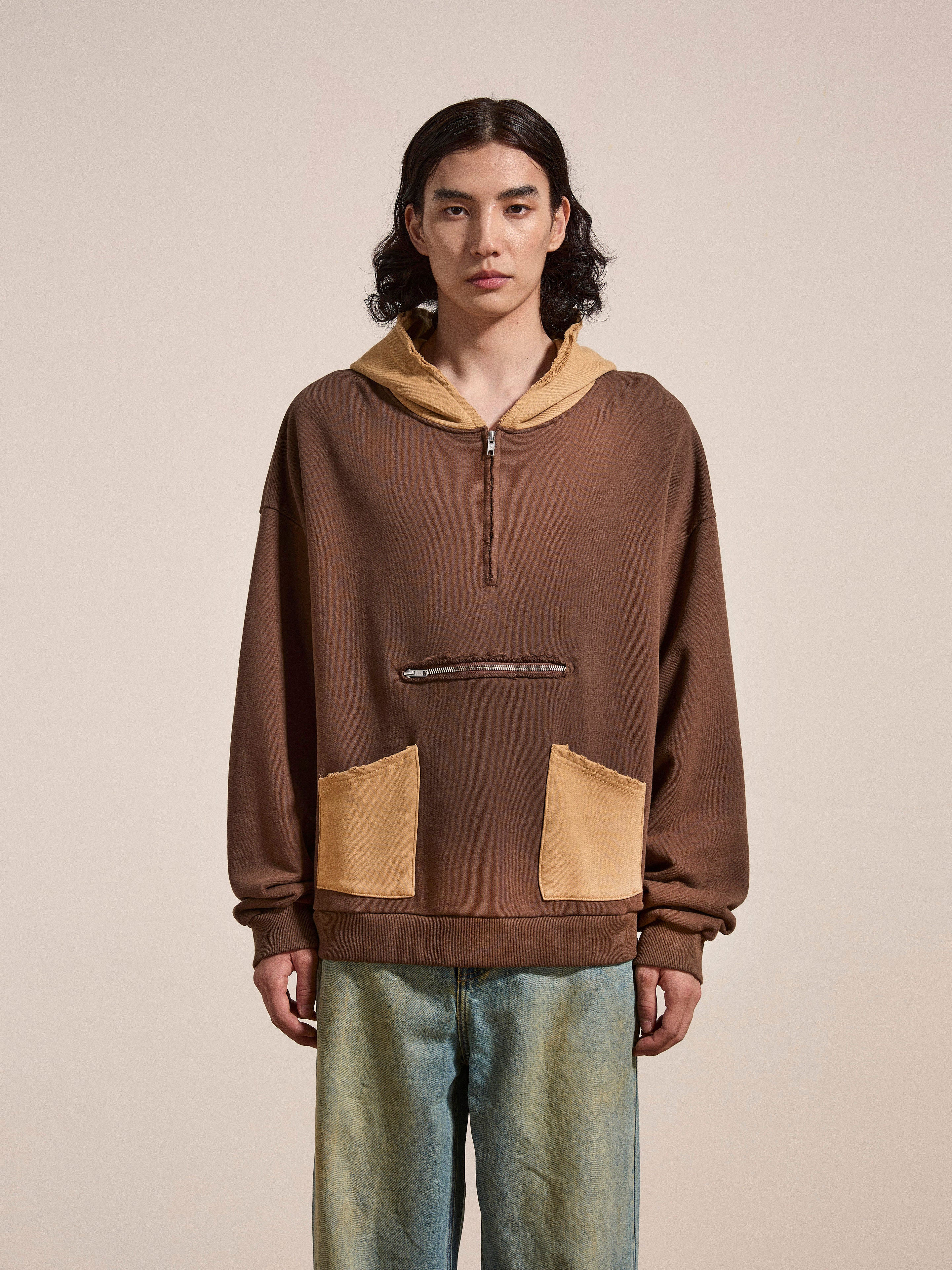 A person with long dark hair stands facing the camera, wearing FOUND's Quarter Zip Two Tone Hoodie in a brown French terry cotton fabric with tan accents, paired with blue jeans. They evoke a vintage feel against a plain beige background.