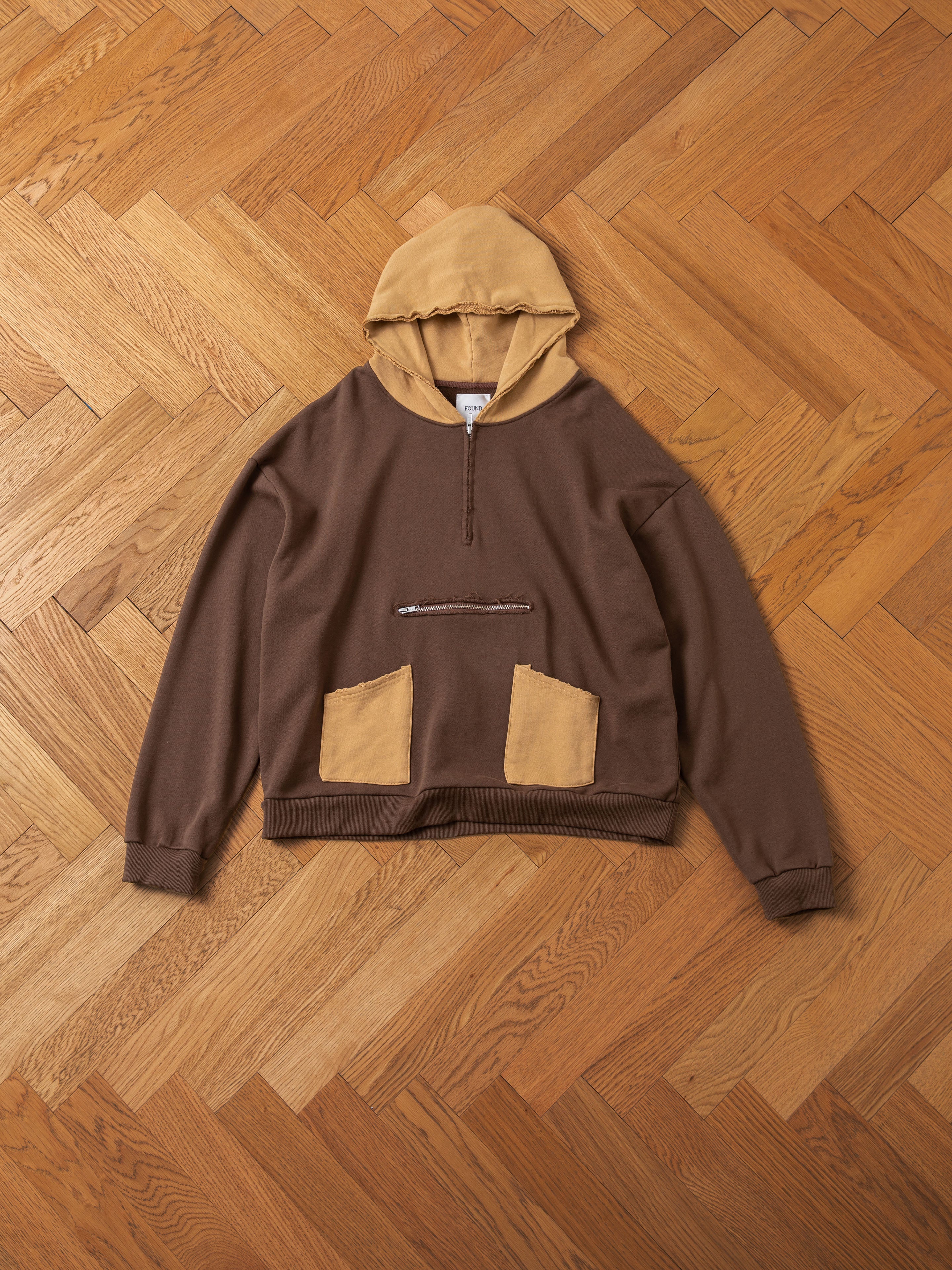 The Quarter Zip Two Tone Hoodie by FOUND, featuring a brown French Terry Cotton body, tan hood and front pockets, and a chest zipper, is lying on a wooden parquet floor.