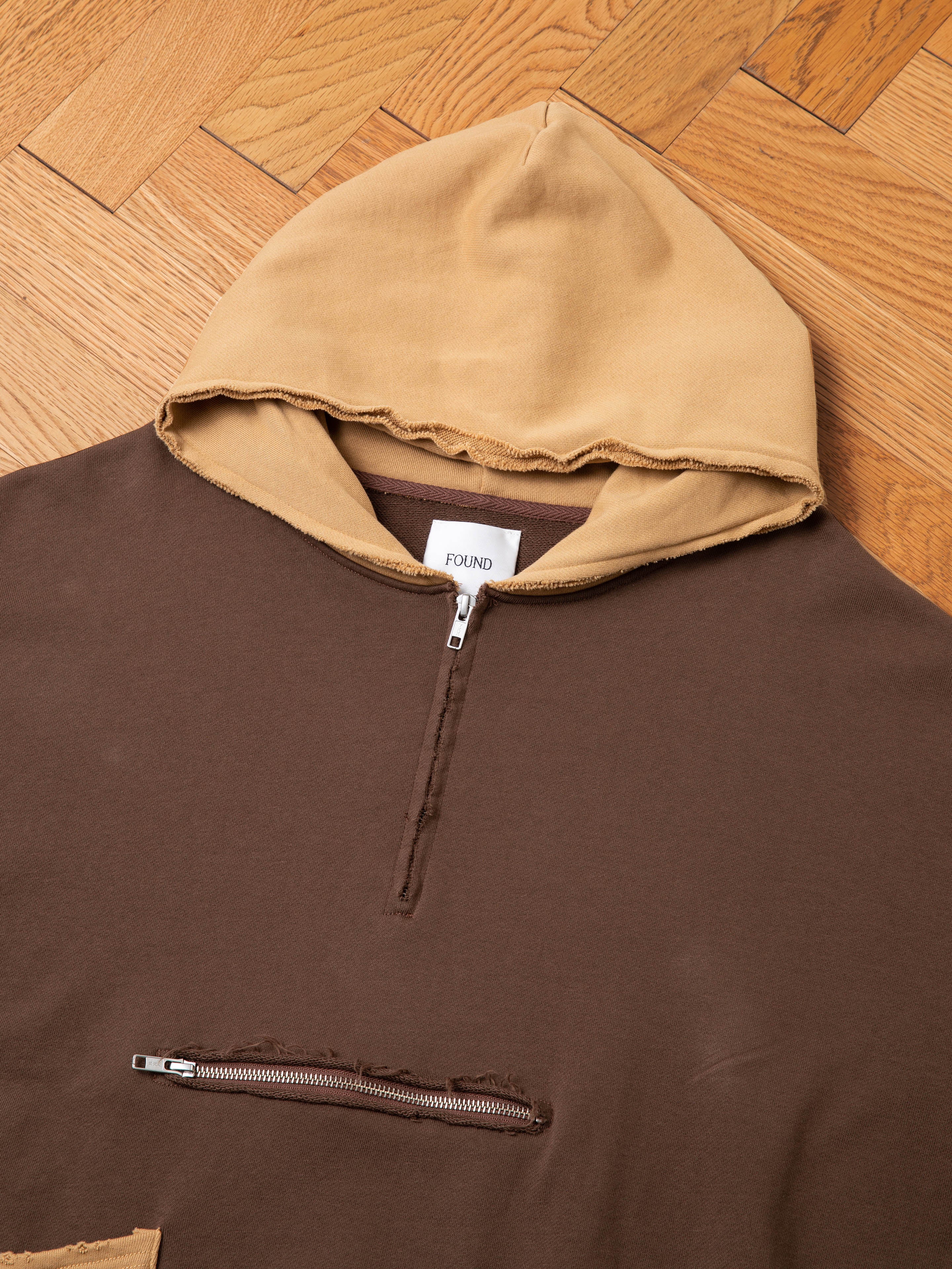 A brown Quarter Zip Two Tone Hoodie with a tan hood, crafted from French Terry Cotton for a vintage feel. It features the brand "FOUND" on a white label and an additional horizontal zipper pocket on the chest. The garment is laid on a wooden floor.