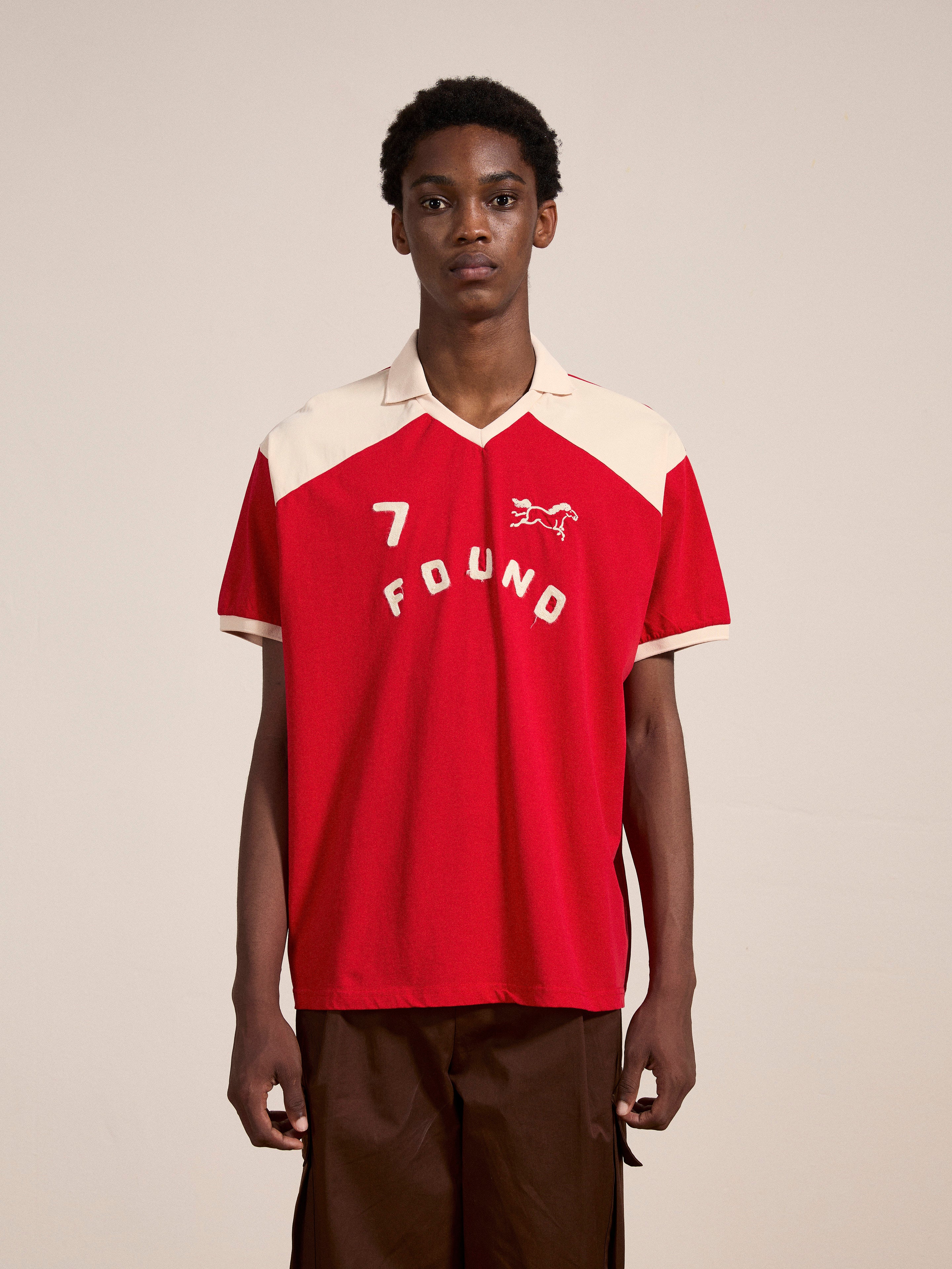 A person stands indoors wearing the Goji Red Jersey by FOUND, featuring the number 7 and the brand name "FOUND" on the front in a style reminiscent of vintage athletic uniforms, complemented by brown pants.