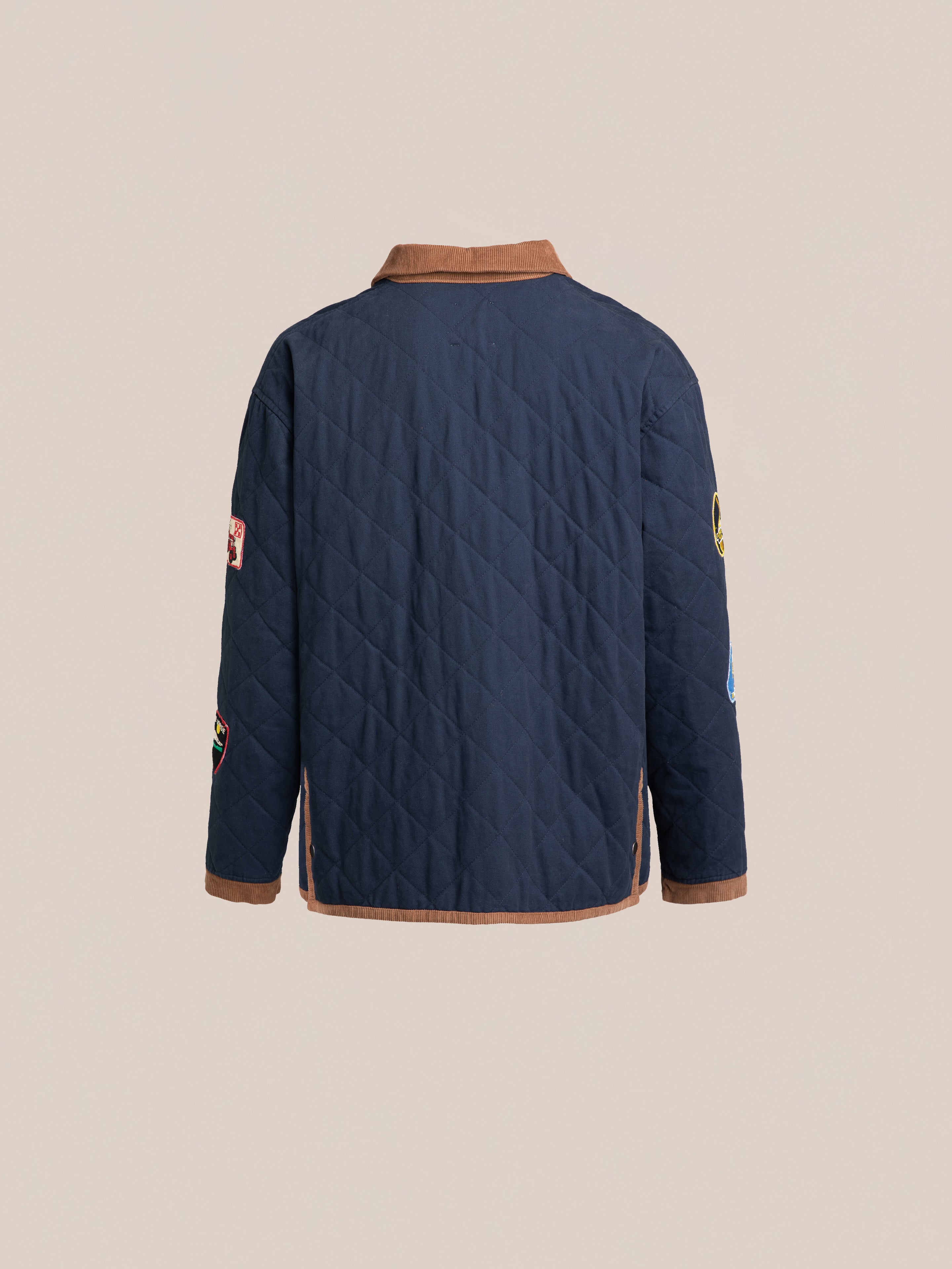 The FOUND Farmstead Quilt Patch Jacket, featuring a navy blue quilted design with a brown collar and sleeve patches that evoke the look of vintage hunting competition jackets, is photographed from the back against a beige background.