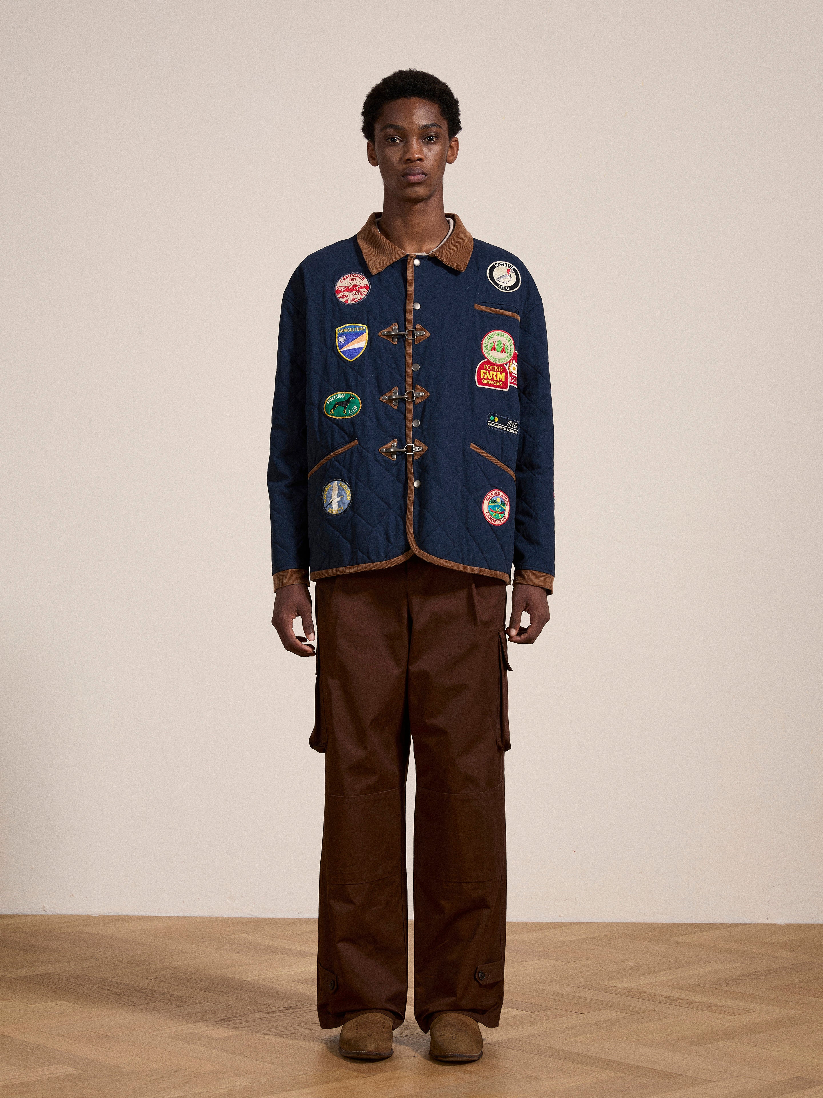 A person stands indoors wearing the FOUND Farmstead Quilt Patch Jacket, a quilted design decorated with various patches reminiscent of vintage hunting competition jackets. They pair it with brown cargo pants and matching shoes.