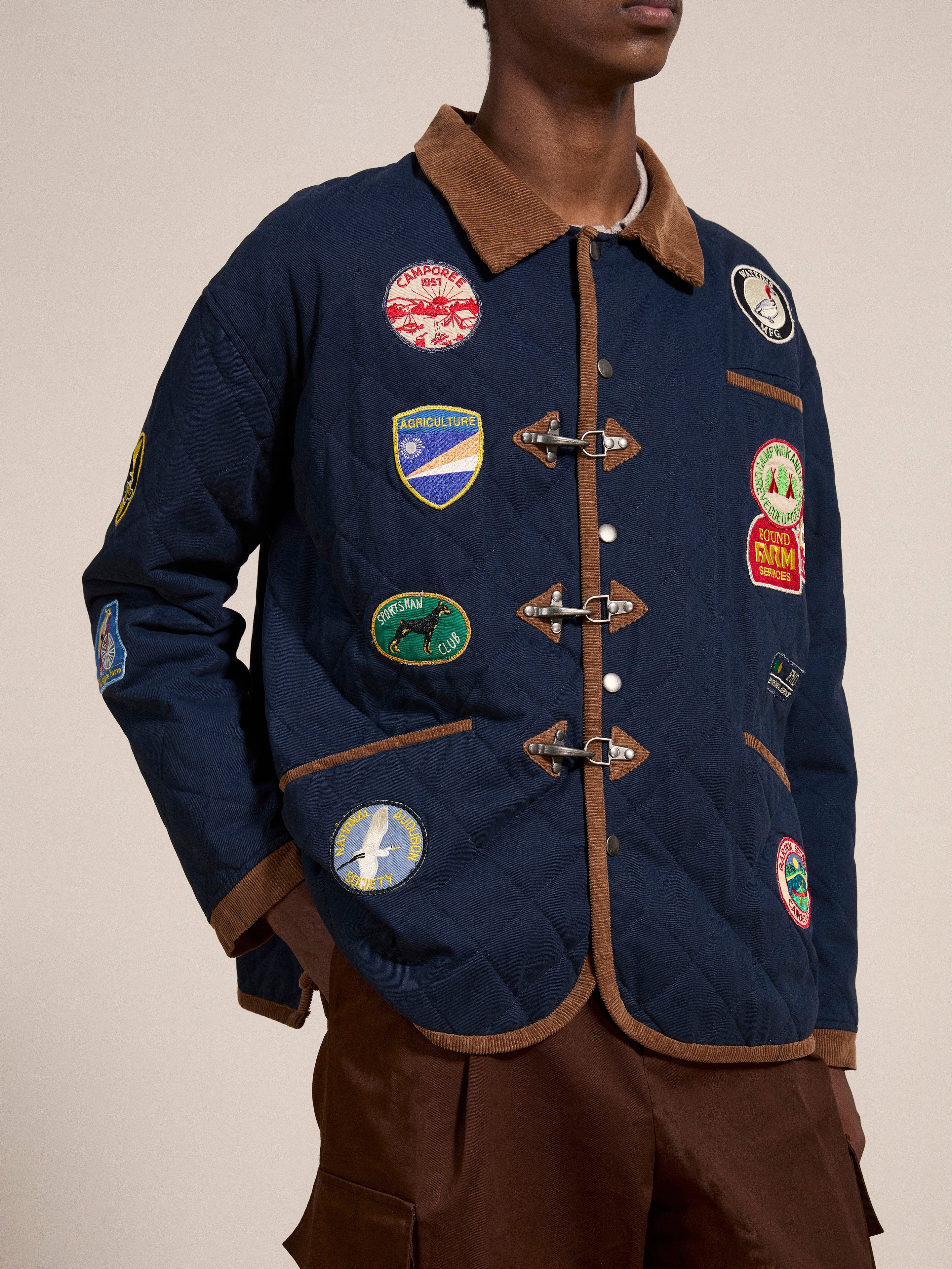 A person is wearing a FOUND Farmstead Quilt Patch Jacket, featuring a quilted navy blue design with vibrant patches and brown accents on the collar, cuffs, and pocket trims. Reminiscent of vintage hunting competition jackets, it also boasts three toggle closures.