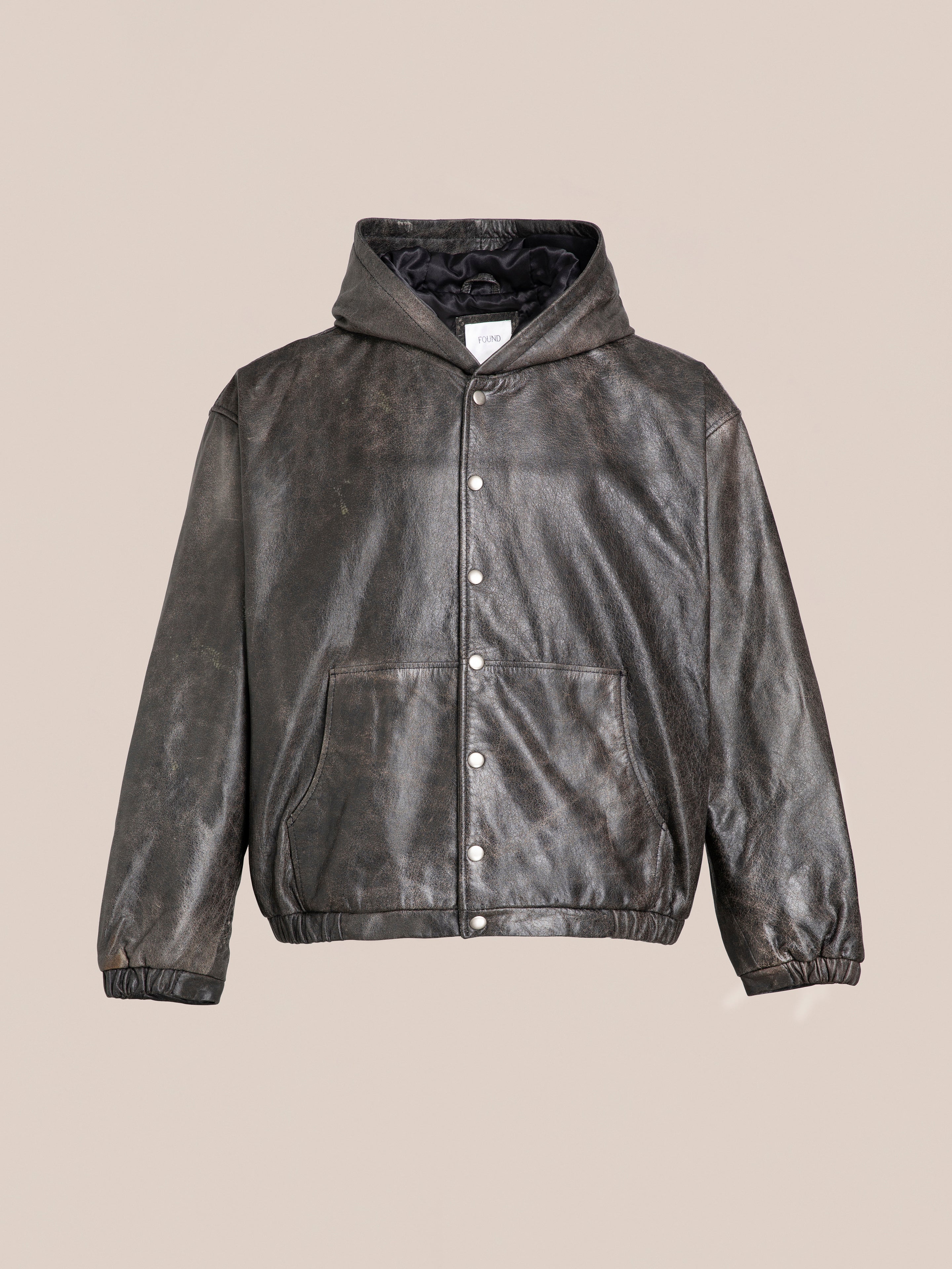 A dark brown Distressed Genuine Leather Hoodie by Found, featuring a hood, front buttons, two pockets, and elastic cuffs, is displayed against a plain beige background.