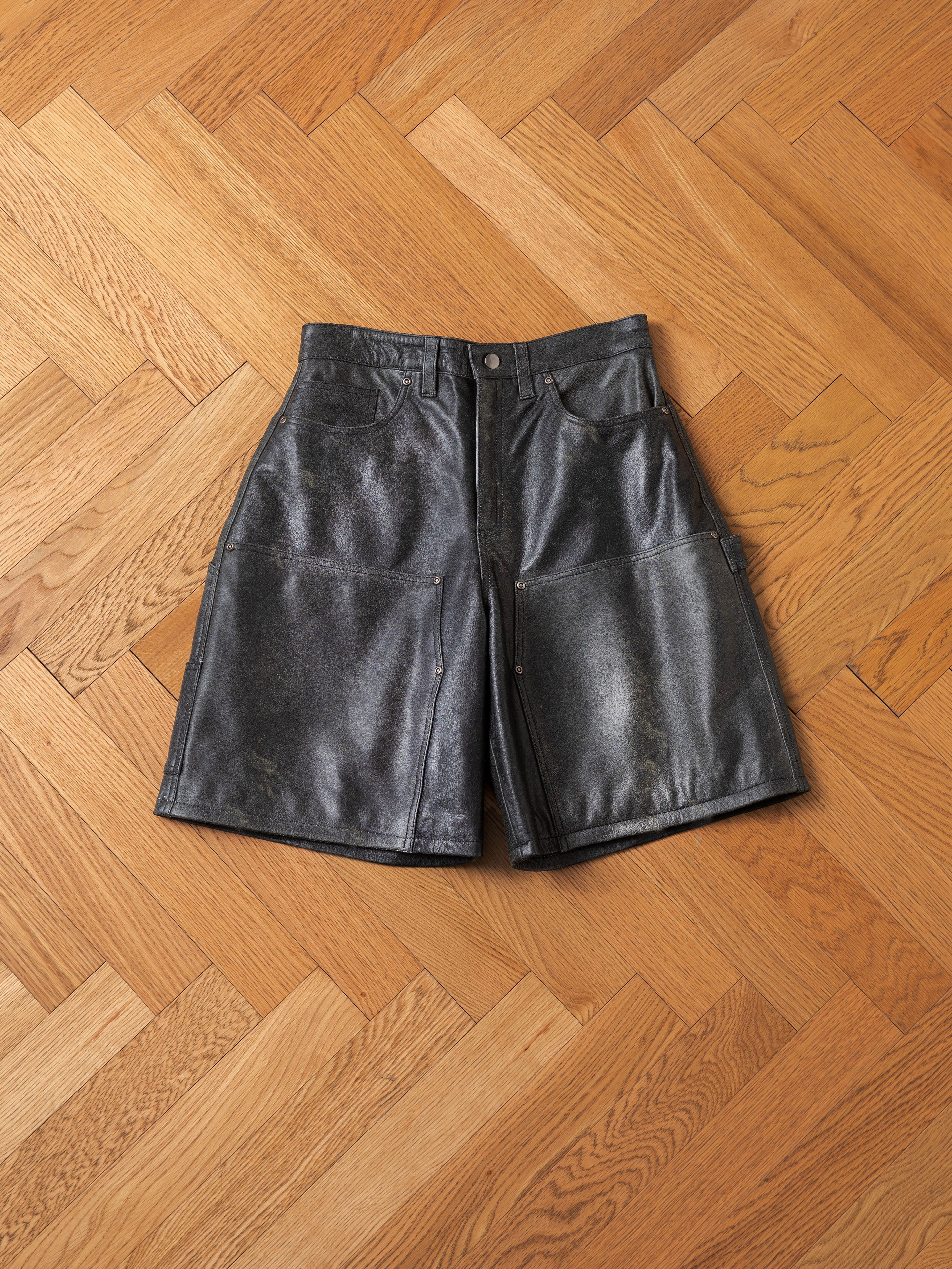 The Distressed Genuine Leather Carpenter Shorts by Found, featuring a double-knee design and front pockets, lay flat on a wooden parquet floor.