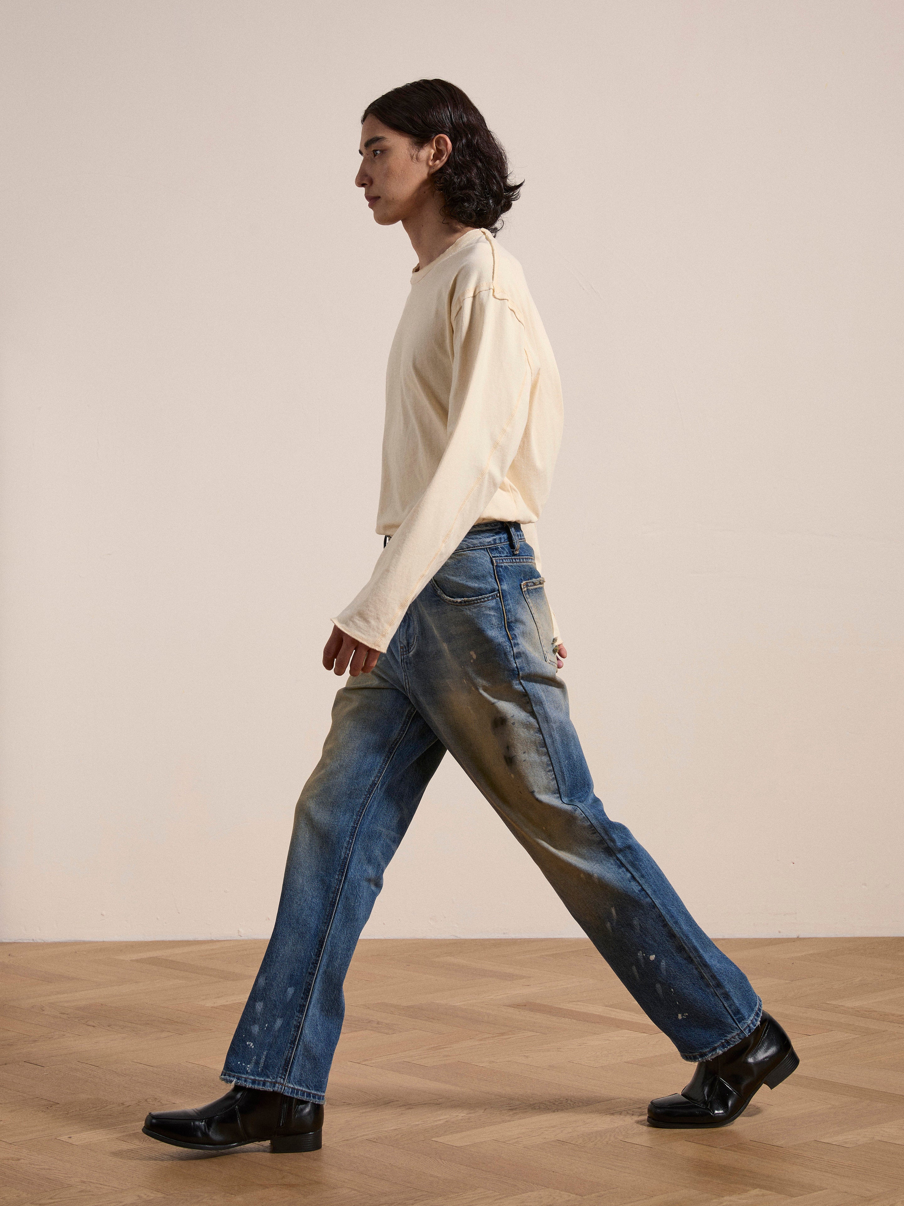 A person with shoulder-length hair walks to the right. They are wearing a long-sleeve beige shirt, FOUND Monet Paint Jeans in a vintage blue wash, and black shoes. The background is a plain, light-colored wall and wooden floor.