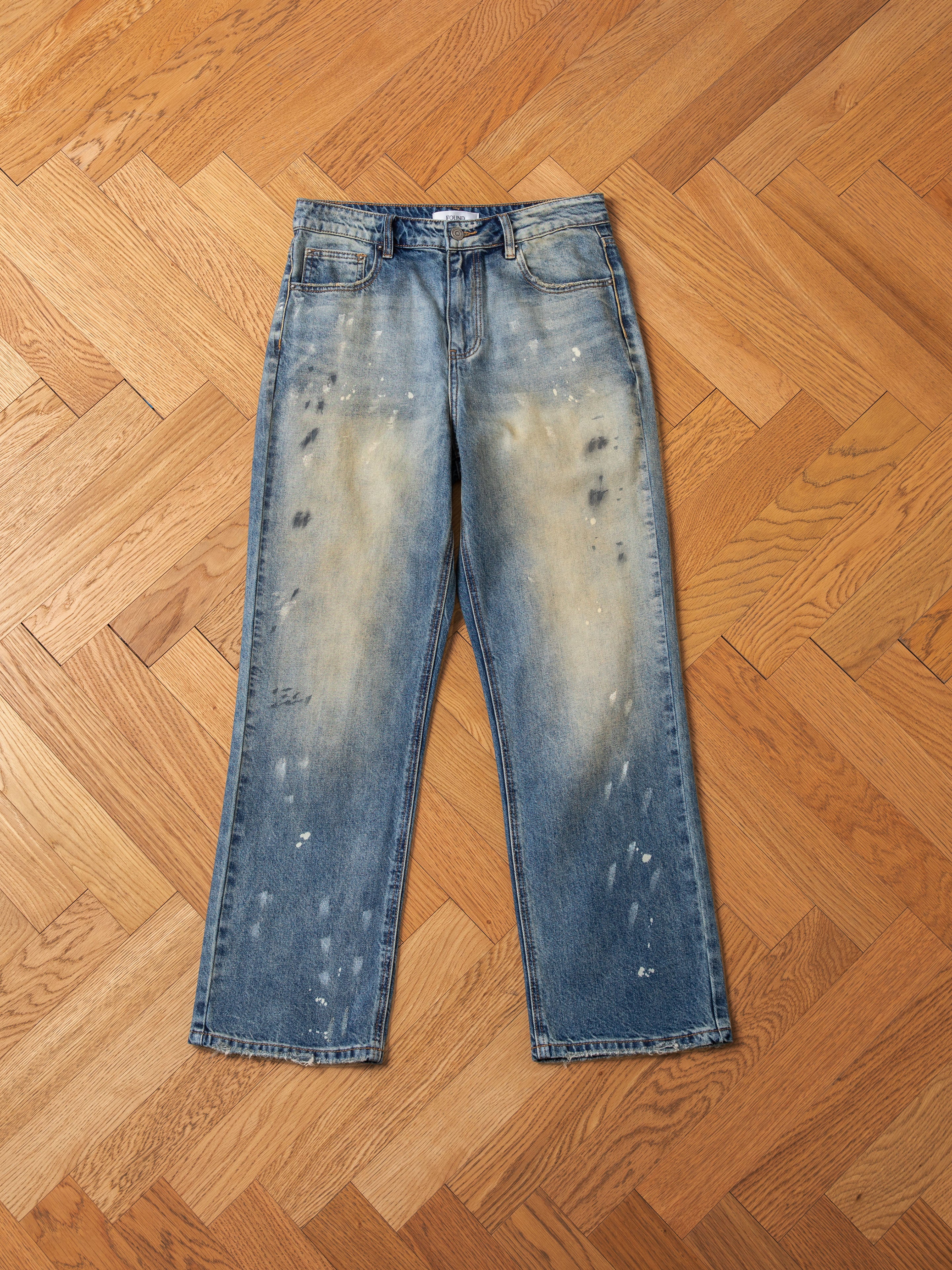 A pair of FOUND Monet Paint Jeans in a vintage blue wash, featuring distressed patches and hand-painted splatter details, laid flat on a herringbone wooden floor.