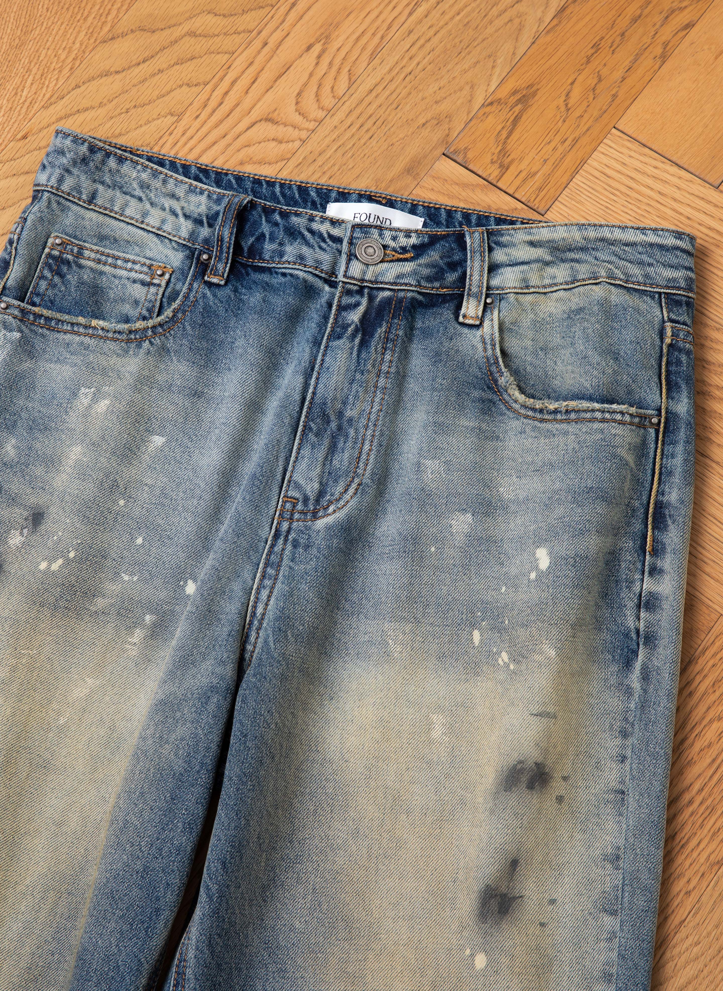 A pair of Monet Paint Jeans by FOUND, showcasing a vintage blue wash with hand-painted splatters and fading details, laid flat on a wooden floor.