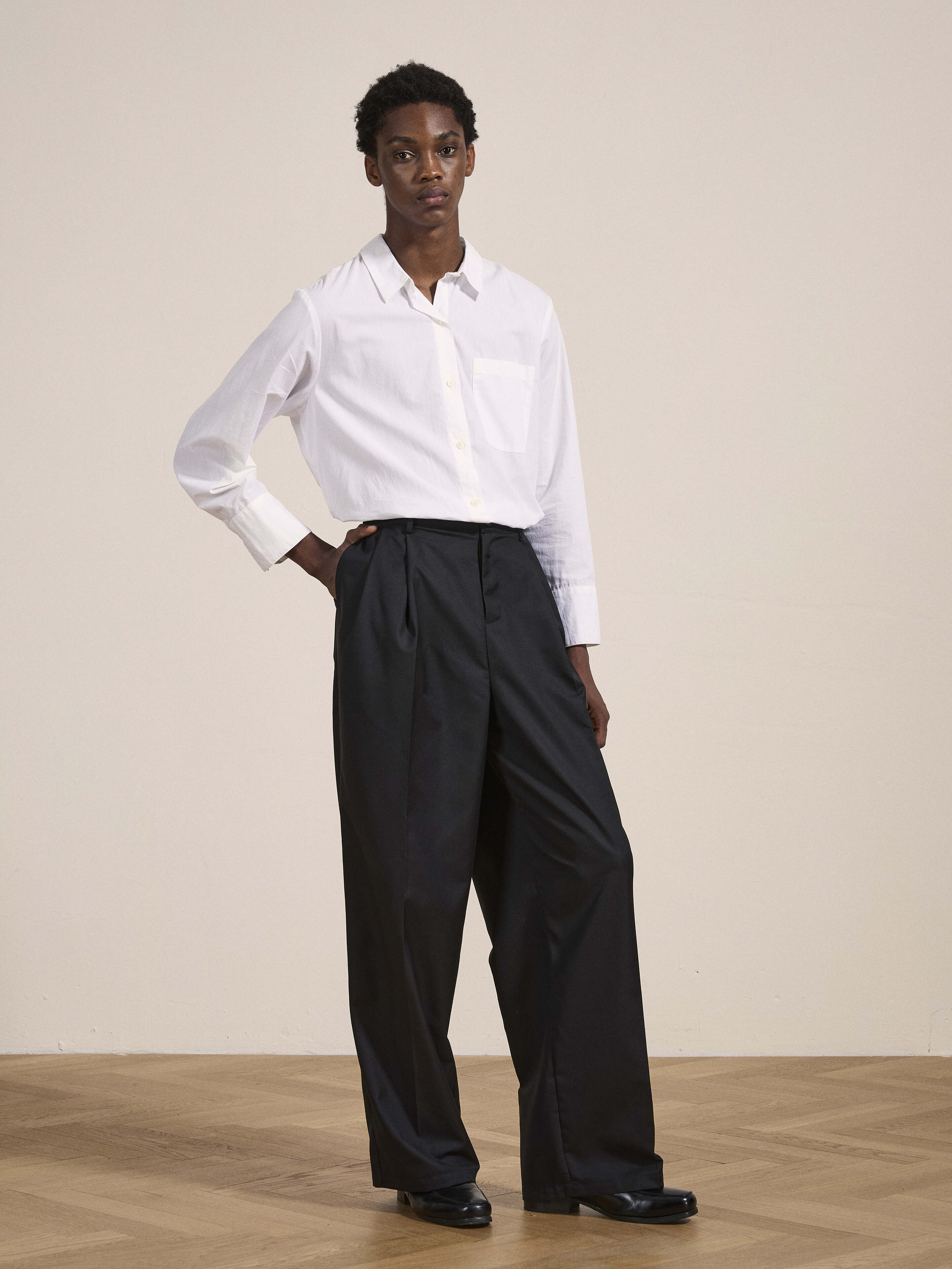 A person stands against a plain background wearing a white shirt and Found's Pleated Trousers, which feature black wide-leg, double pleats, and adjustable waist tabs, with one hand resting on their hip.