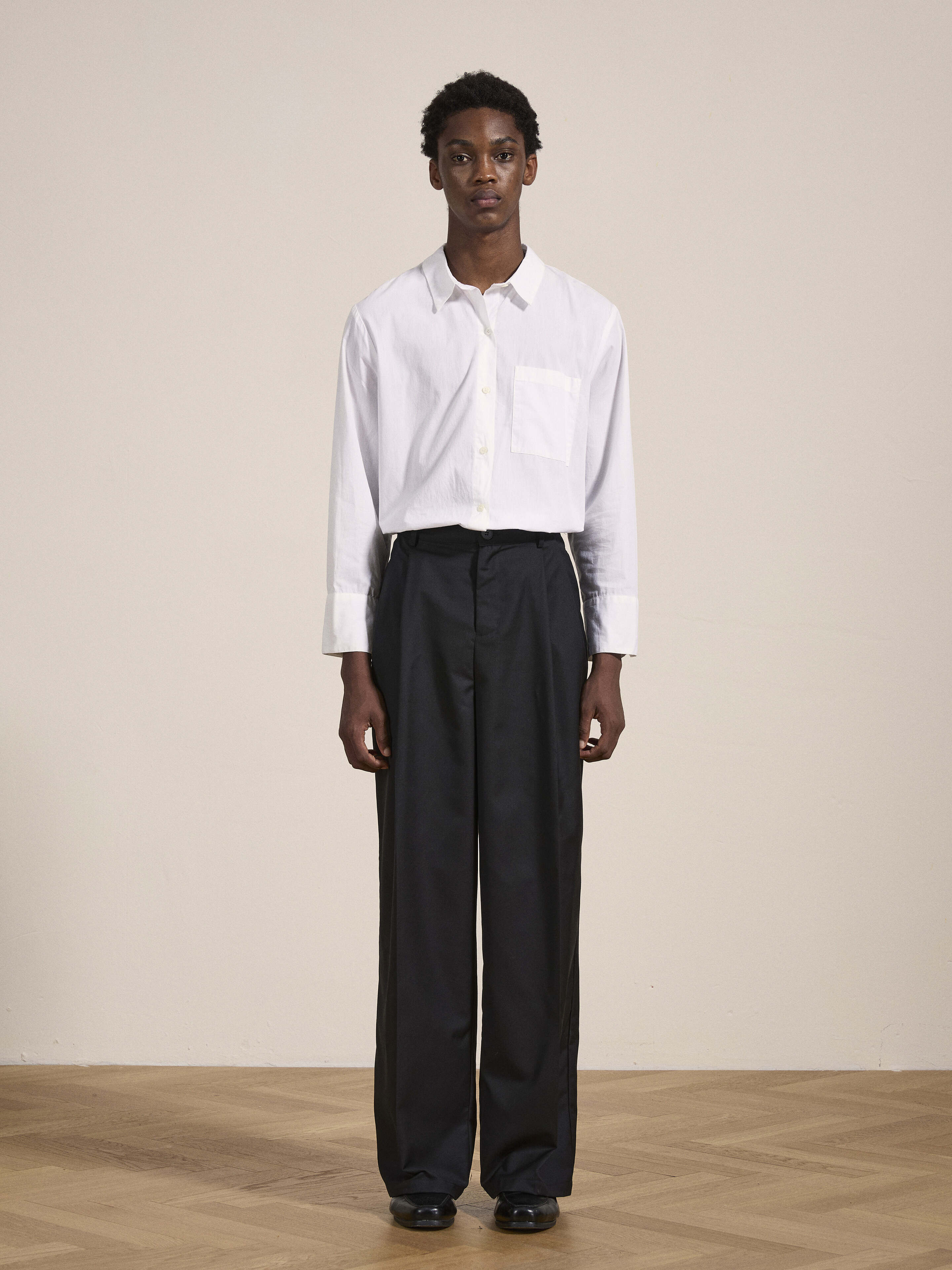 Person standing indoors against a plain background, wearing a white dress shirt and Found Pleated Trousers with adjustable waist tabs, featuring a neutral facial expression.