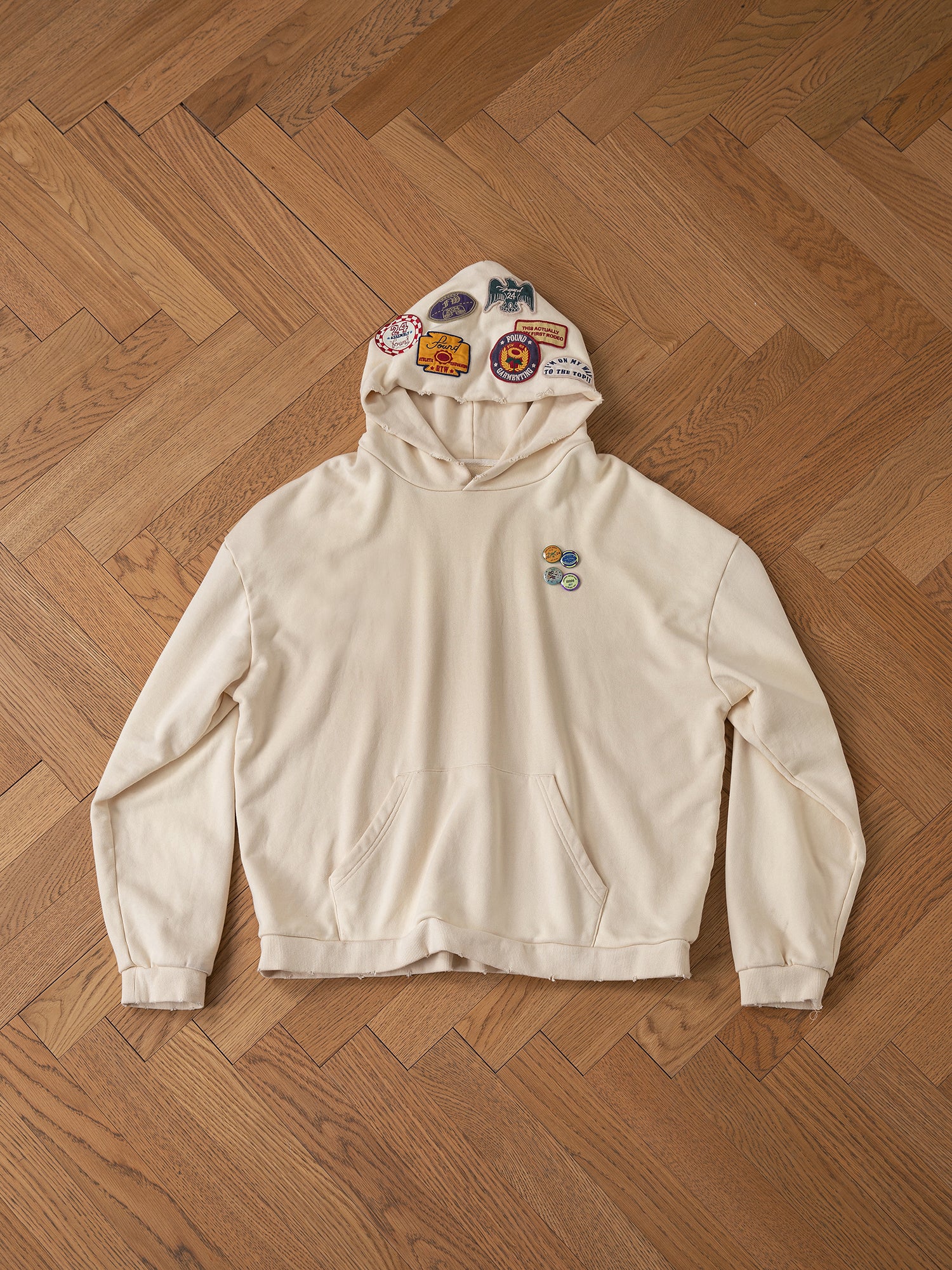 FOUND's Faded Patch Hoodie showcases a vintage flair with its oversized fit, embellished with an array of patches on the hood and chest, elegantly displayed flat on a wooden floor.