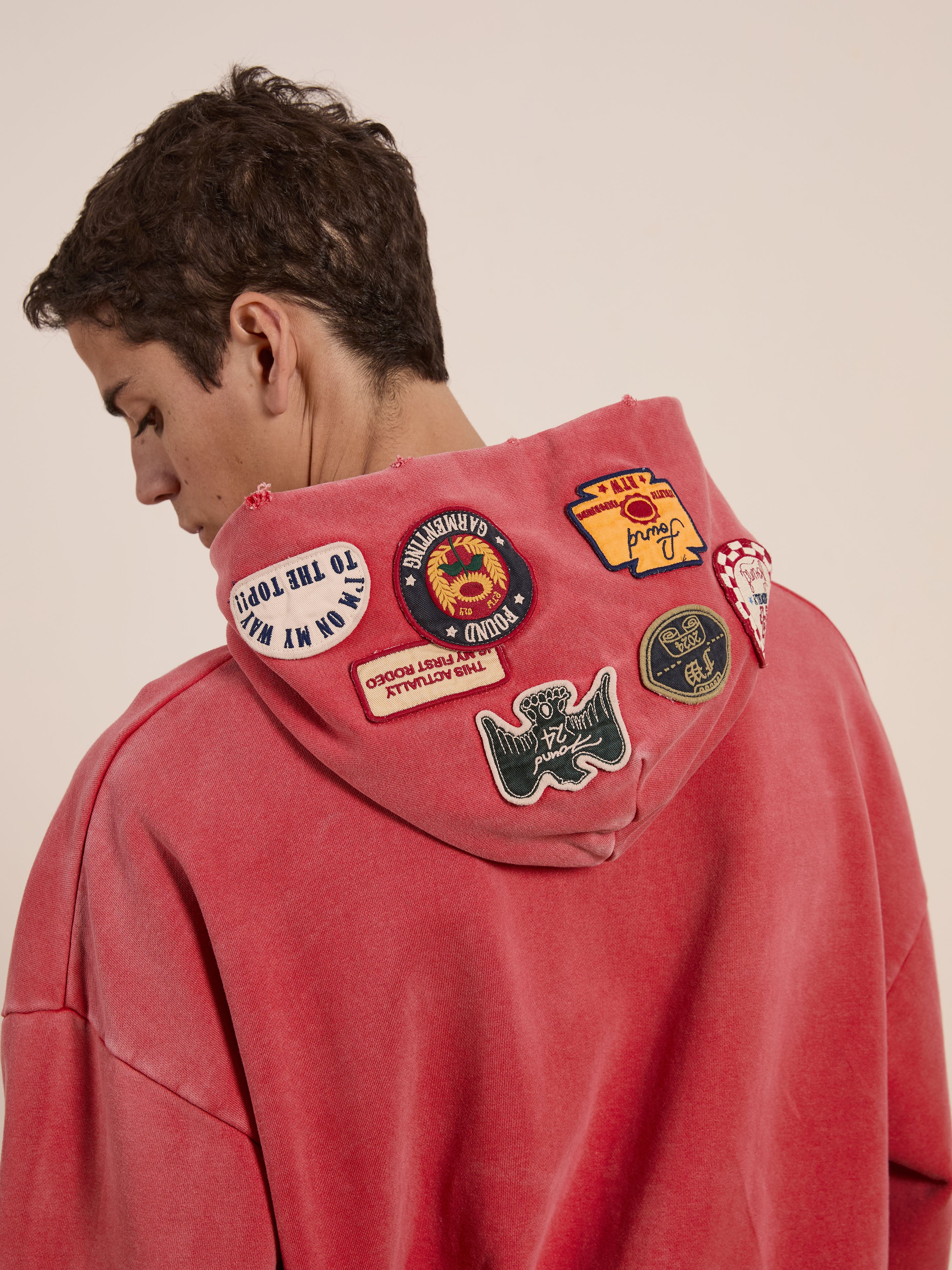 A person wearing FOUND's Multi Patch Hoodie in vintage-inspired red with a sun-faded wash and colorful patches on the back of the hood, facing away.