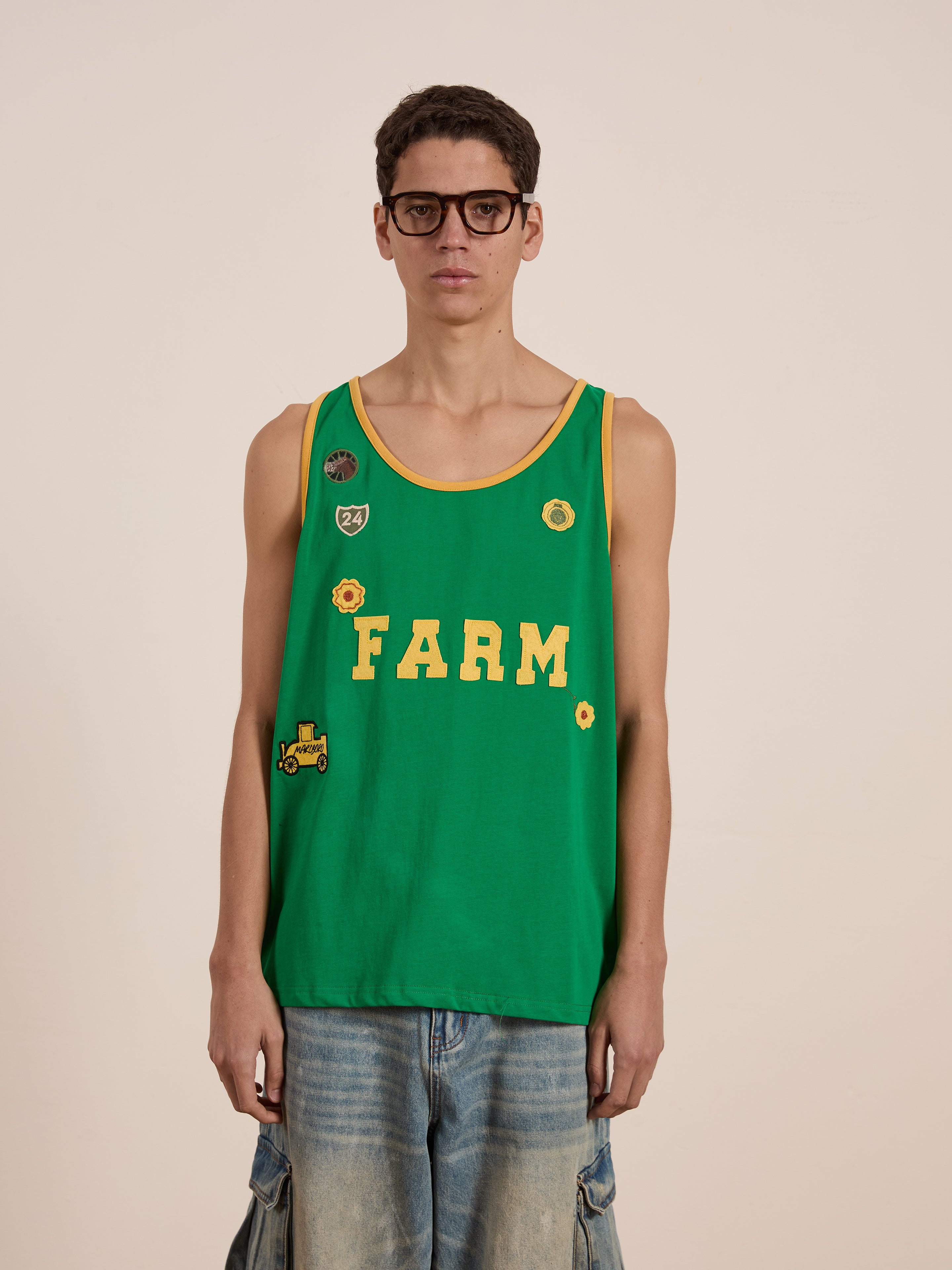 Wearing the "Farm Tank" by FOUND, featuring embroidered patches and paired with light blue jeans, a person showcases style against a neutral background. Made from breathable cotton, the green tank top effortlessly blends comfort and fashion.