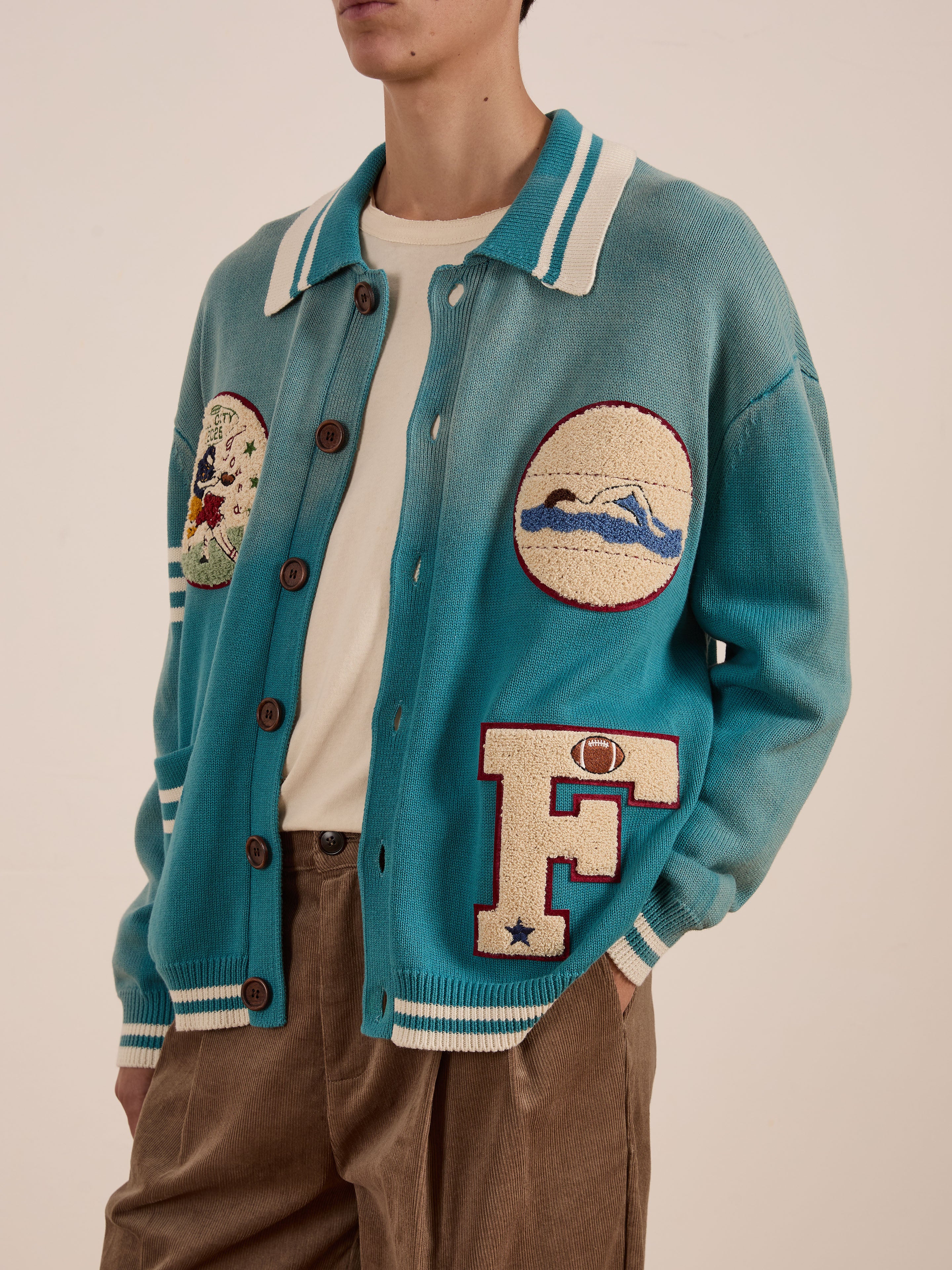 A person features the FOUND Fin Varsity Patch Collared Cardigan in teal, with prominent embroidered patches, over a crisp white shirt and classic brown pants, creating a collegiate style that radiates vintage charm.