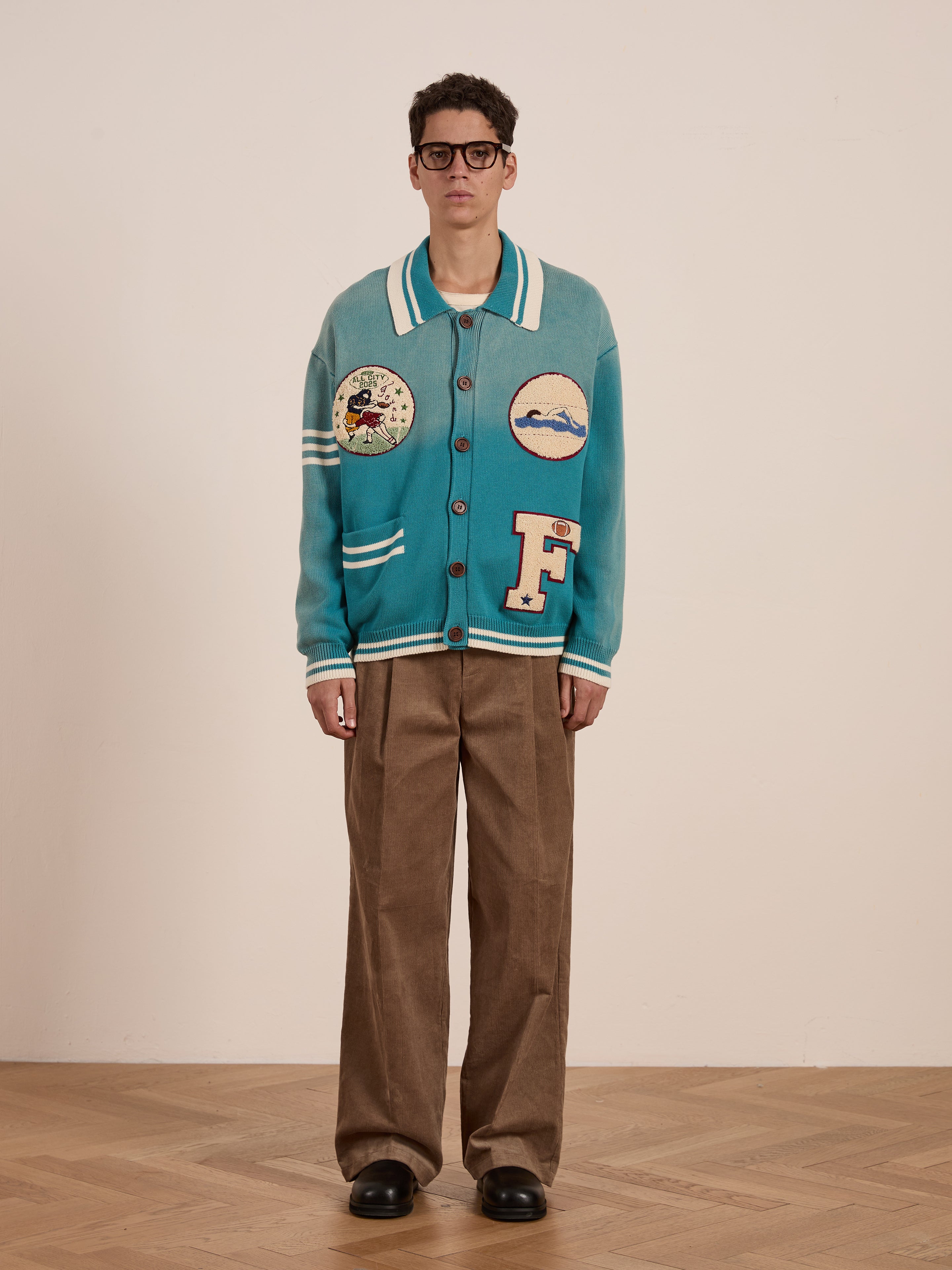 A person in FOUND's Fin Varsity Patch Collared Cardigan, featuring a vintage-inspired teal button-up design with varsity patches, pairs it with brown wide-leg pants and glasses, standing on a wooden floor against a plain backdrop.