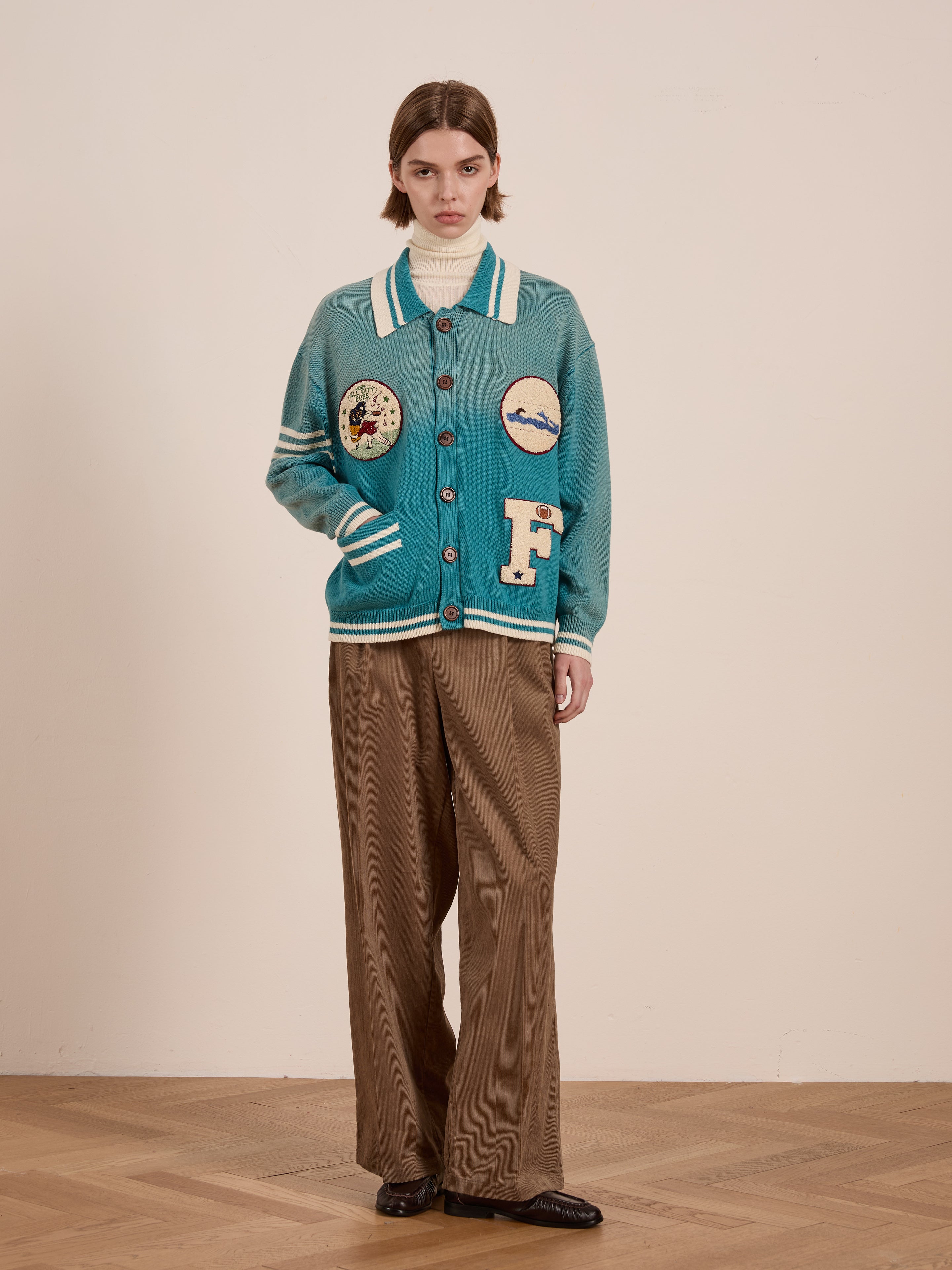 A person showcases a vintage-inspired look wearing FOUND's Fin Varsity Patch Collared Cardigan in blue, paired with a white turtleneck and brown pants. They stand gracefully on a wooden floor against a beige background.