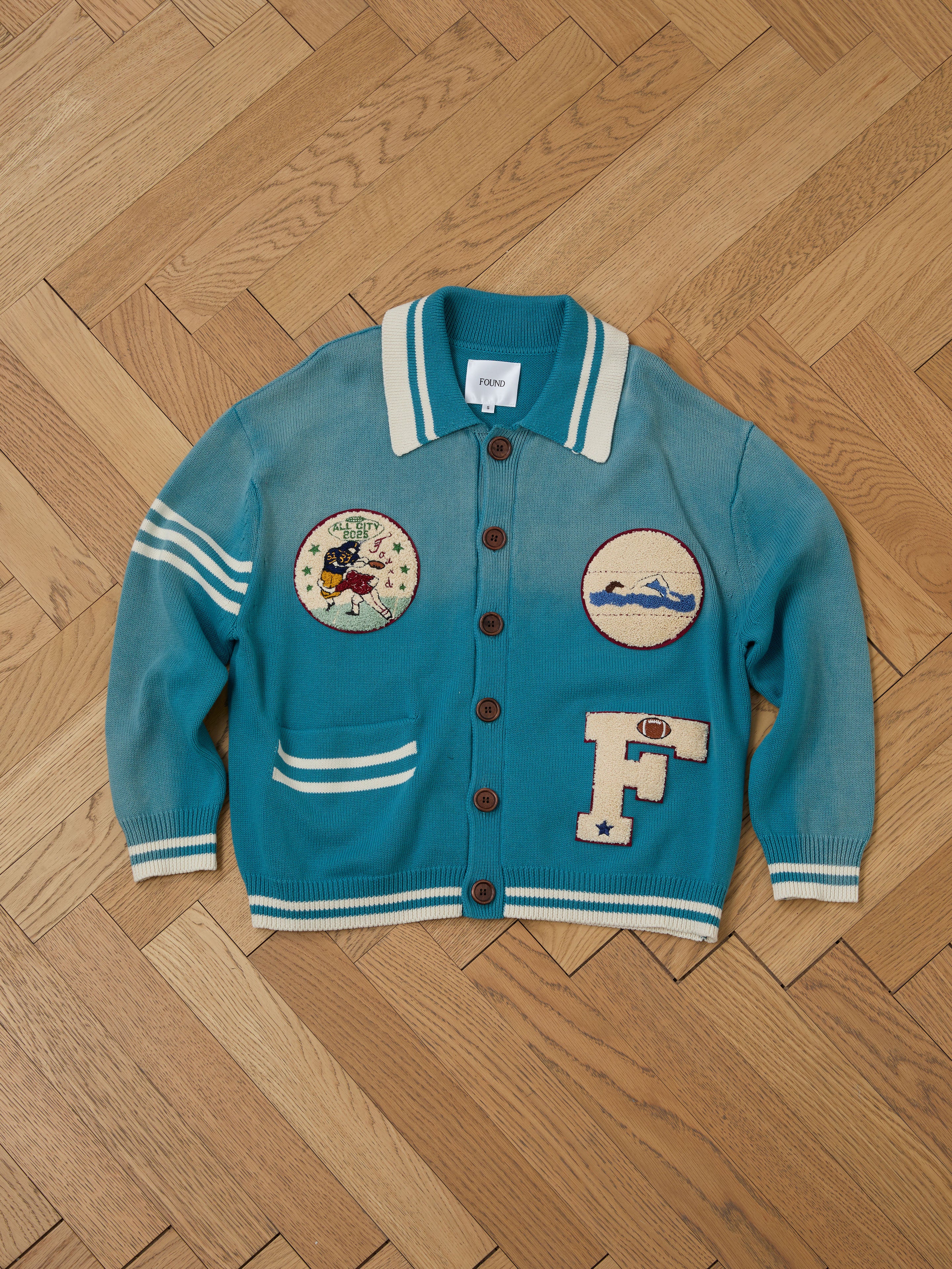 The Fin Varsity Patch Collared Cardigan by FOUND displays a collegiate style on a wooden floor with its striped cuffs and collar. Its button closure reveals vintage-inspired patches, featuring a cartoon figure, mountains, and the letter "F.