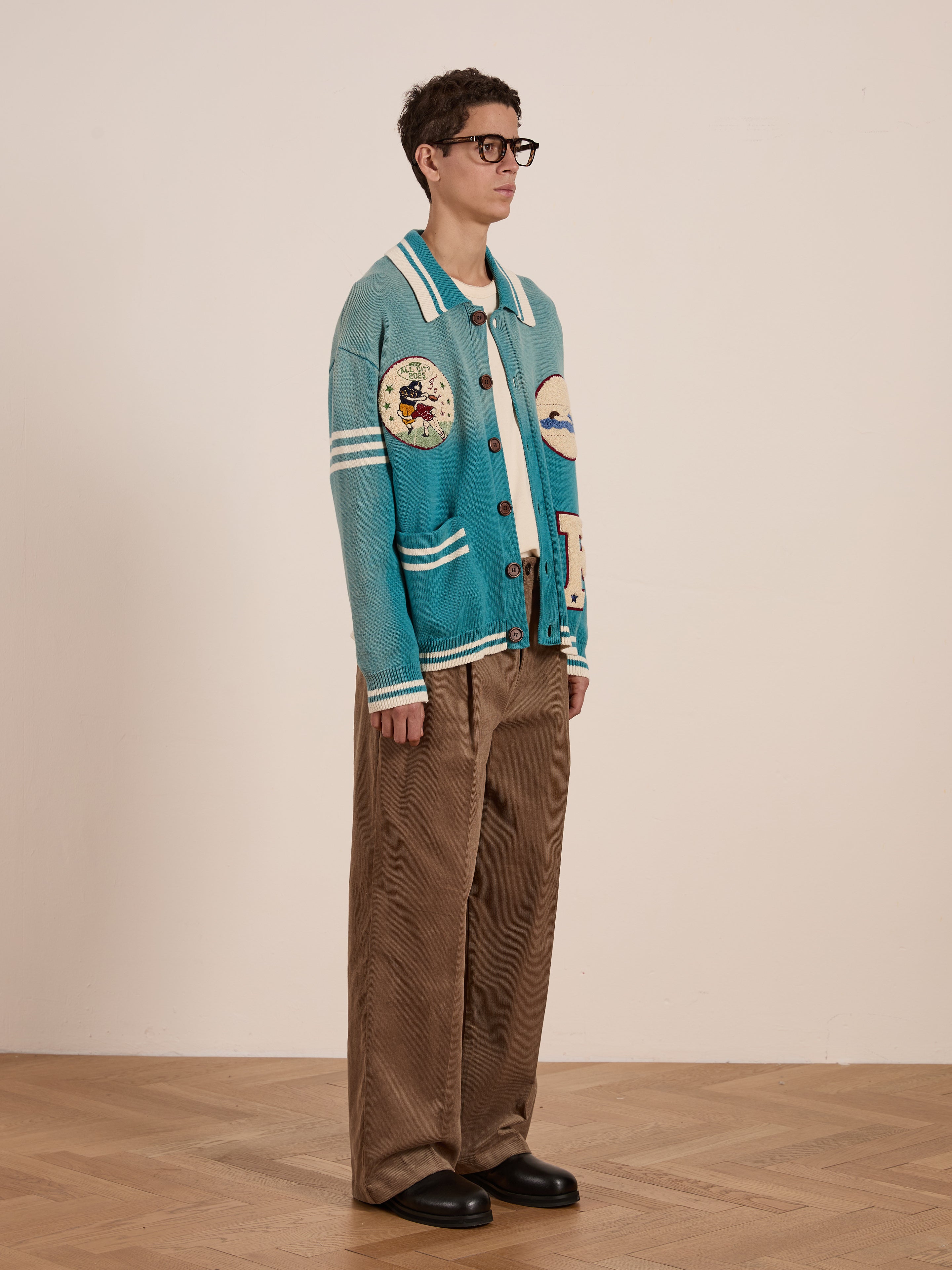 On a wooden floor against a plain wall, a person wears the FOUND Fin Varsity Patch Collared Cardigan with patterns, paired with brown wide-leg pants and glasses, giving a touch of collegiate style to the vintage-inspired look.