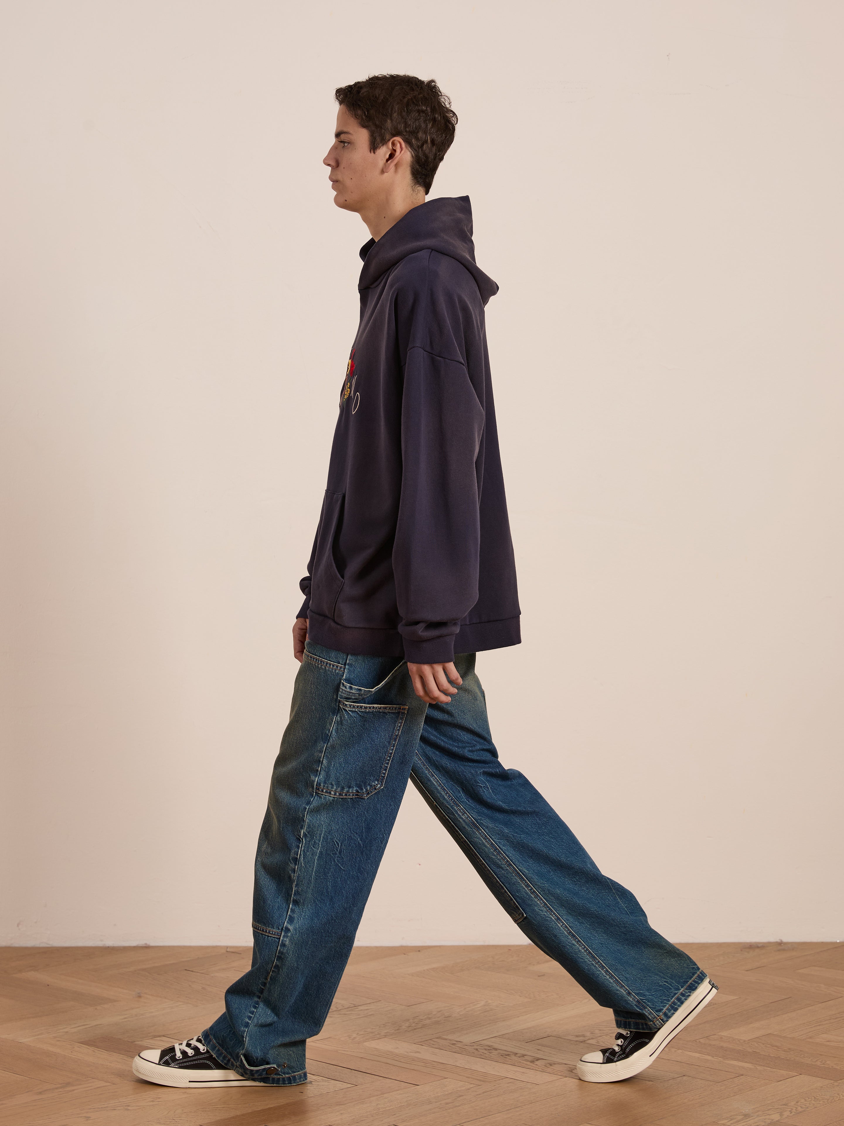 A person in a dark Floral Embroidered Deer Hoodie by FOUND and wide-leg jeans walks on a wooden floor, facing left.