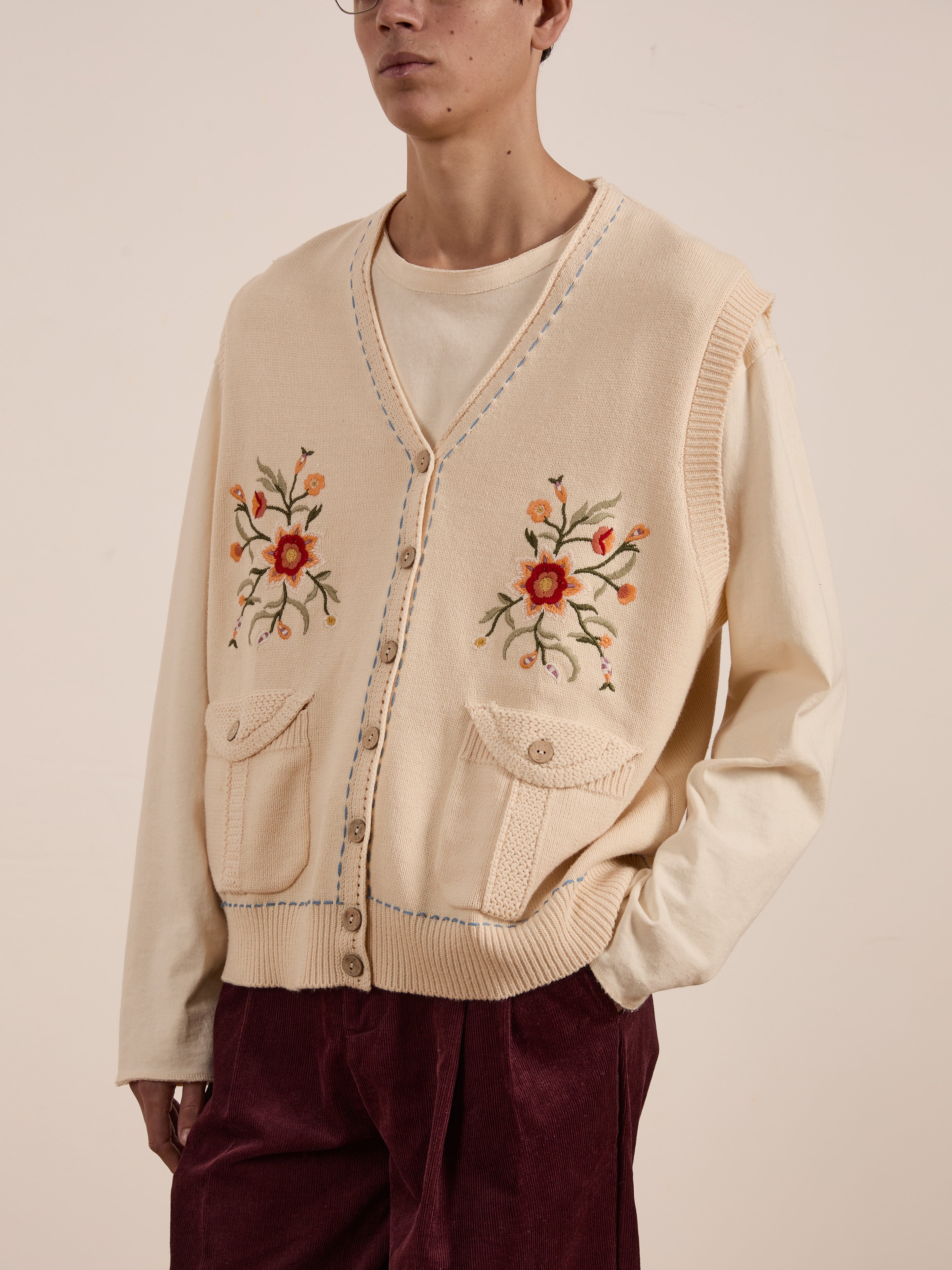 A person is wearing the Floral Embroidered Knit Vest by FOUND, a vintage-inspired cream-colored sleeveless knit with floral embroidery, over a beige shirt and maroon pants.