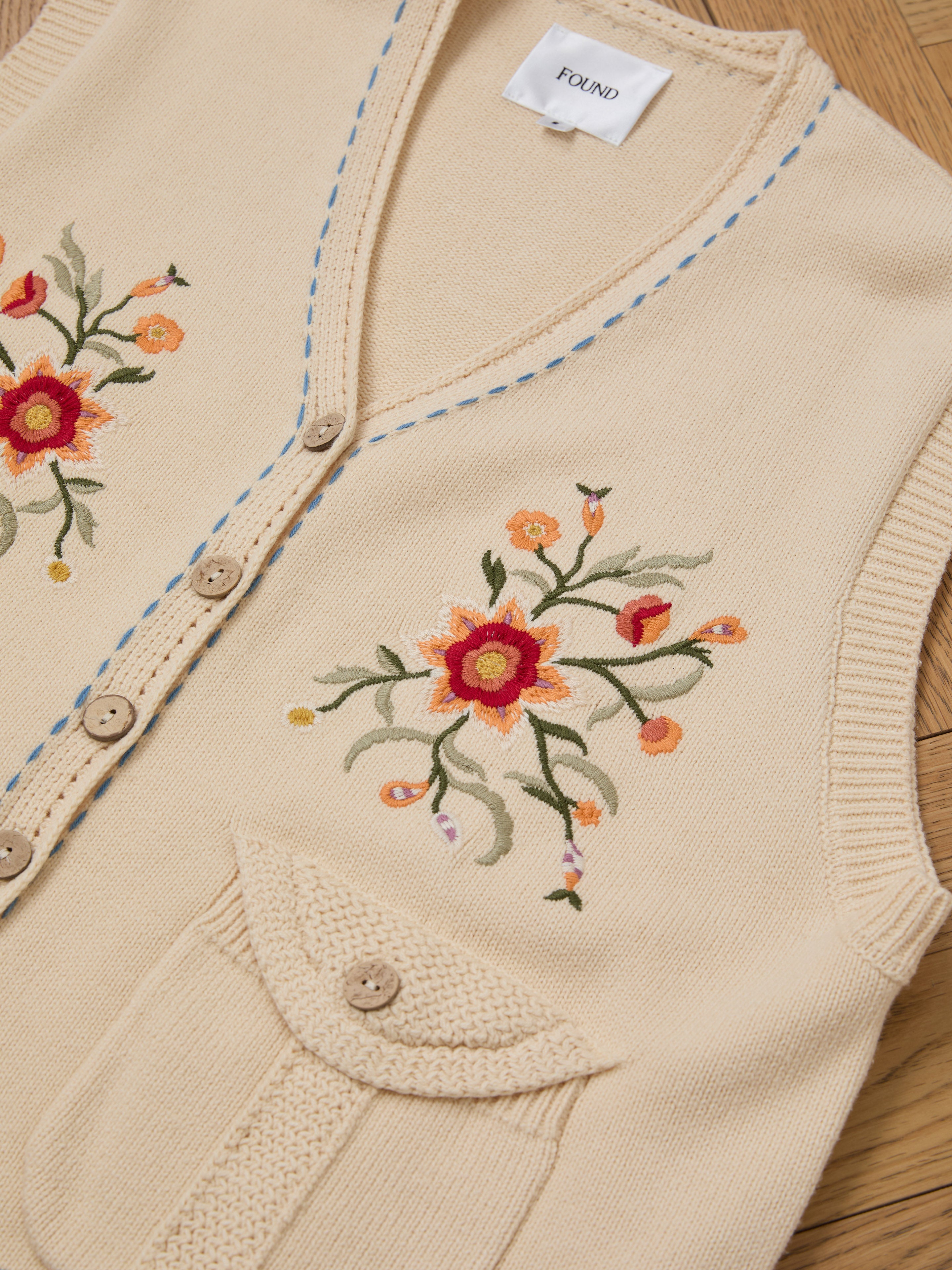 The Floral Embroidered Knit Vest by FOUND is a vintage-inspired beige piece with exquisite floral designs, wooden buttons, and a charming flap pocket. It's perfect for unisex styling and adds timeless elegance when displayed on a wooden surface.