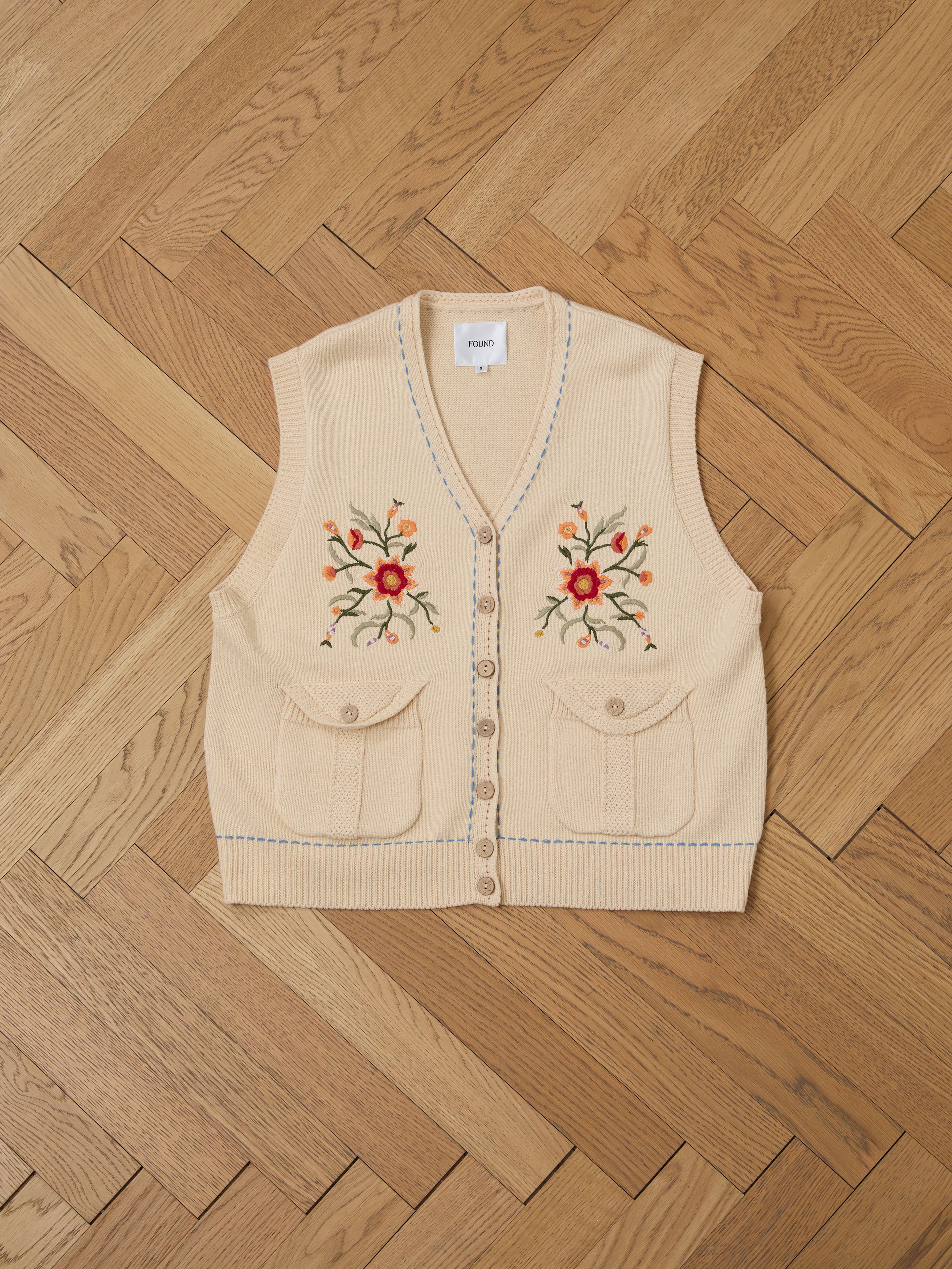 The Floral Embroidered Knit Vest by FOUND, a vintage-inspired beige knit vest with front buttons and two pockets adorned with colorful floral embroidery, is elegantly displayed on a wooden herringbone floor.