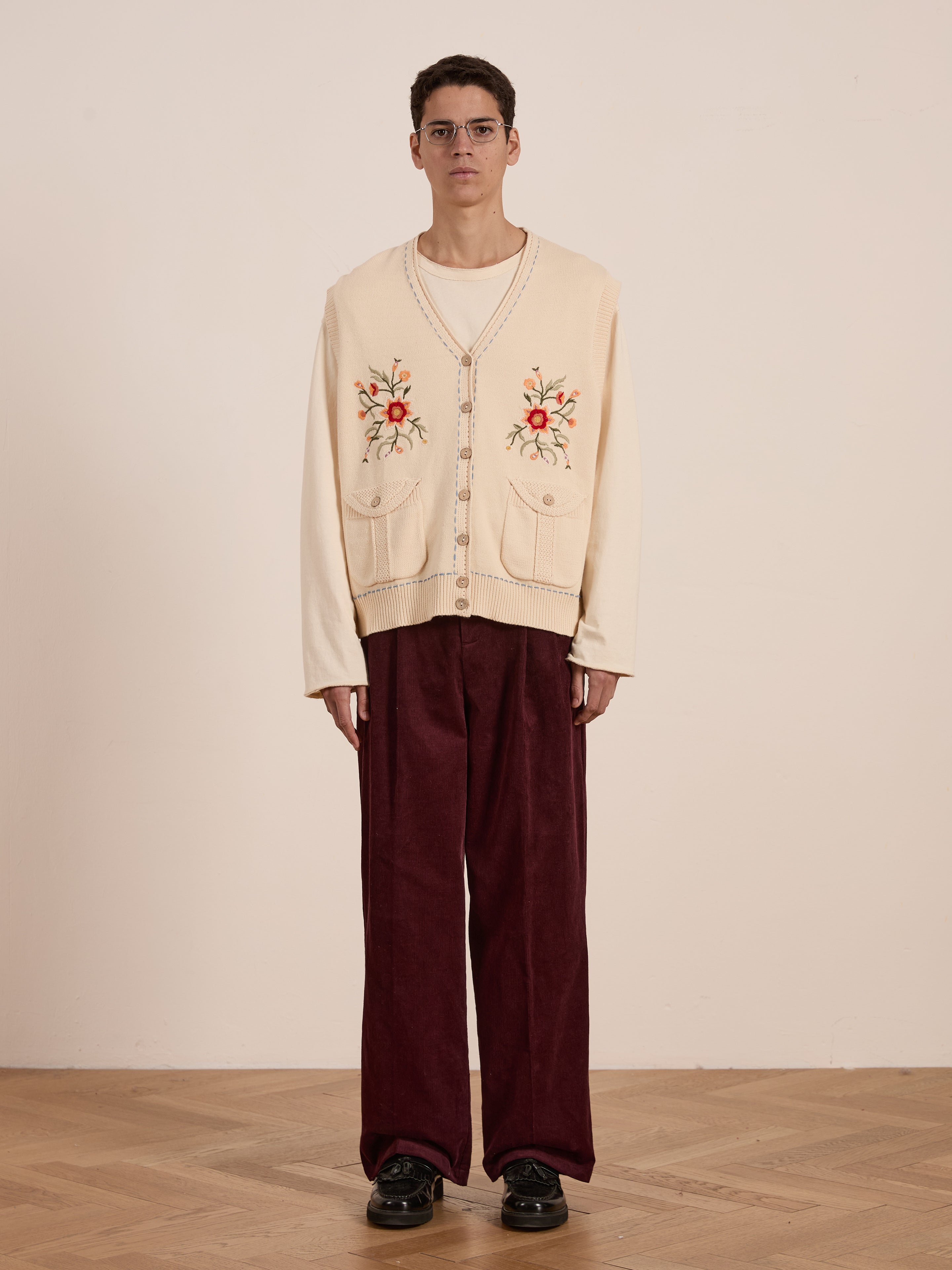 A person wearing glasses, a vintage-inspired Floral Embroidered Knit Vest by FOUND in beige, and burgundy pants stands on a wooden floor against a plain background.