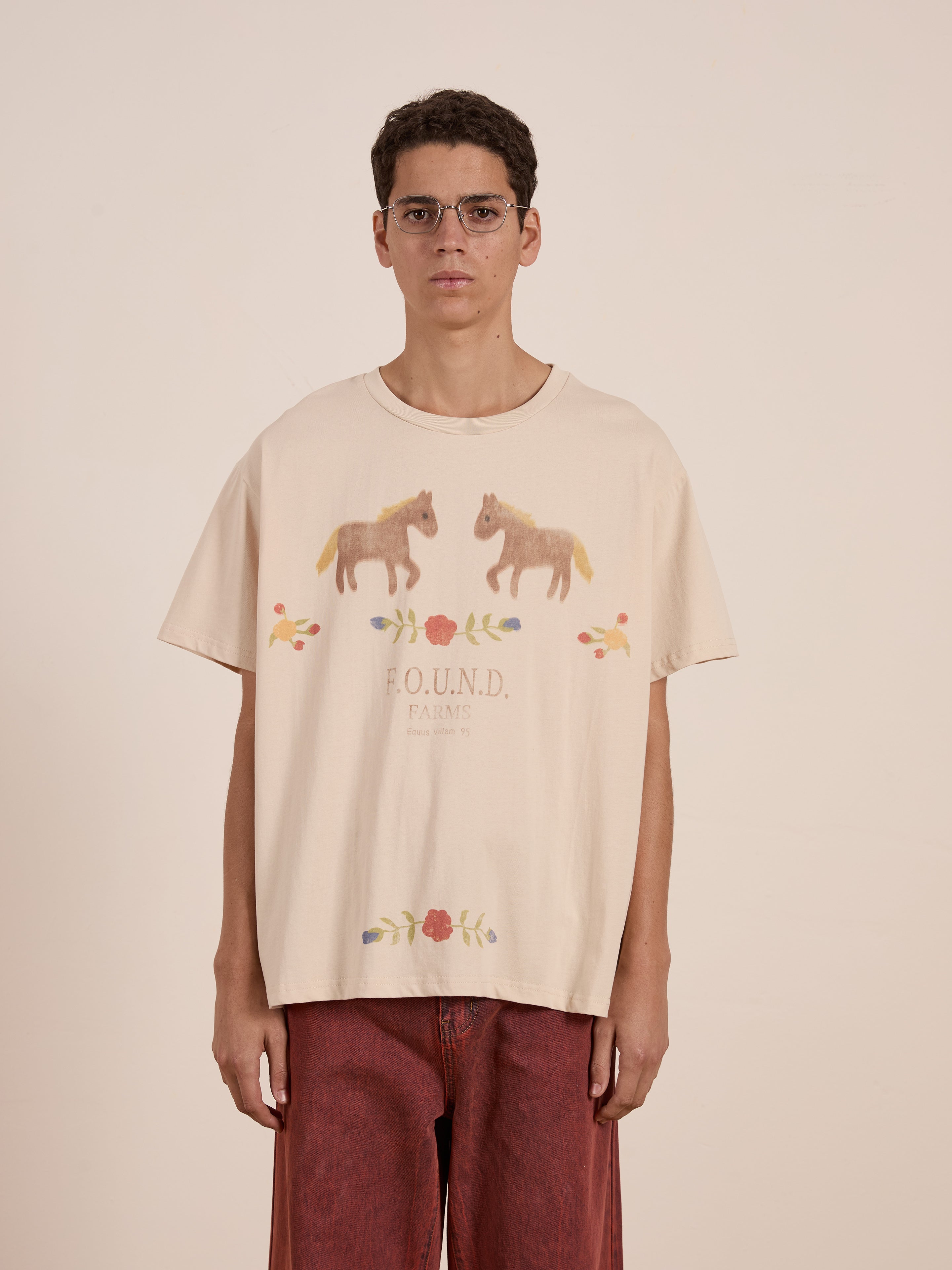 A person in glasses wears a FOUND Floral Horse Farm Tee, showcasing equestrian and floral designs against a plain backdrop.