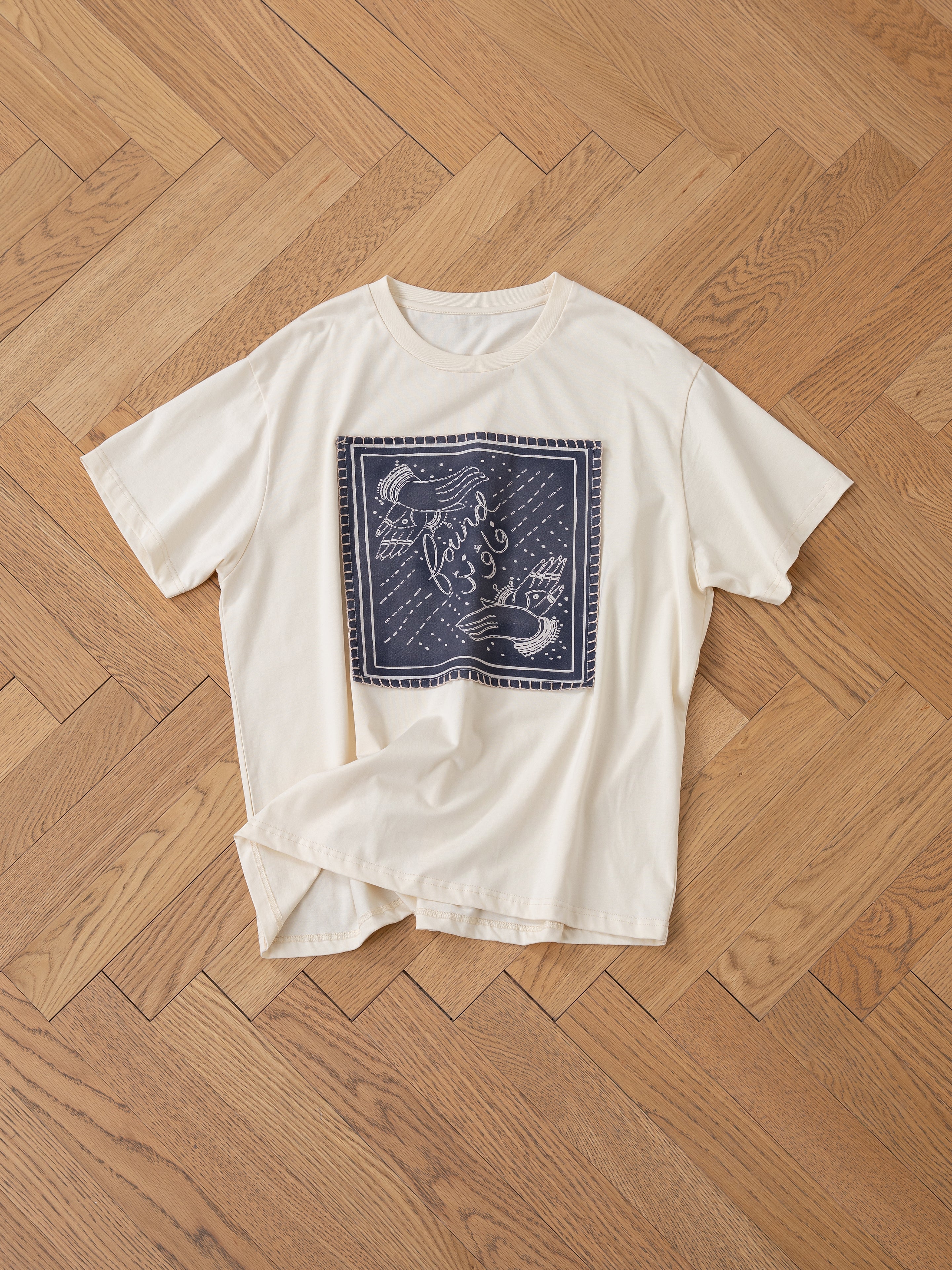 The Hand Art Graphic Tee by FOUND, a cotton cream t-shirt featuring a dark blue square graphic inspired by Mughal paintings with bird motifs and text, is elegantly laid on a wooden herringbone floor.