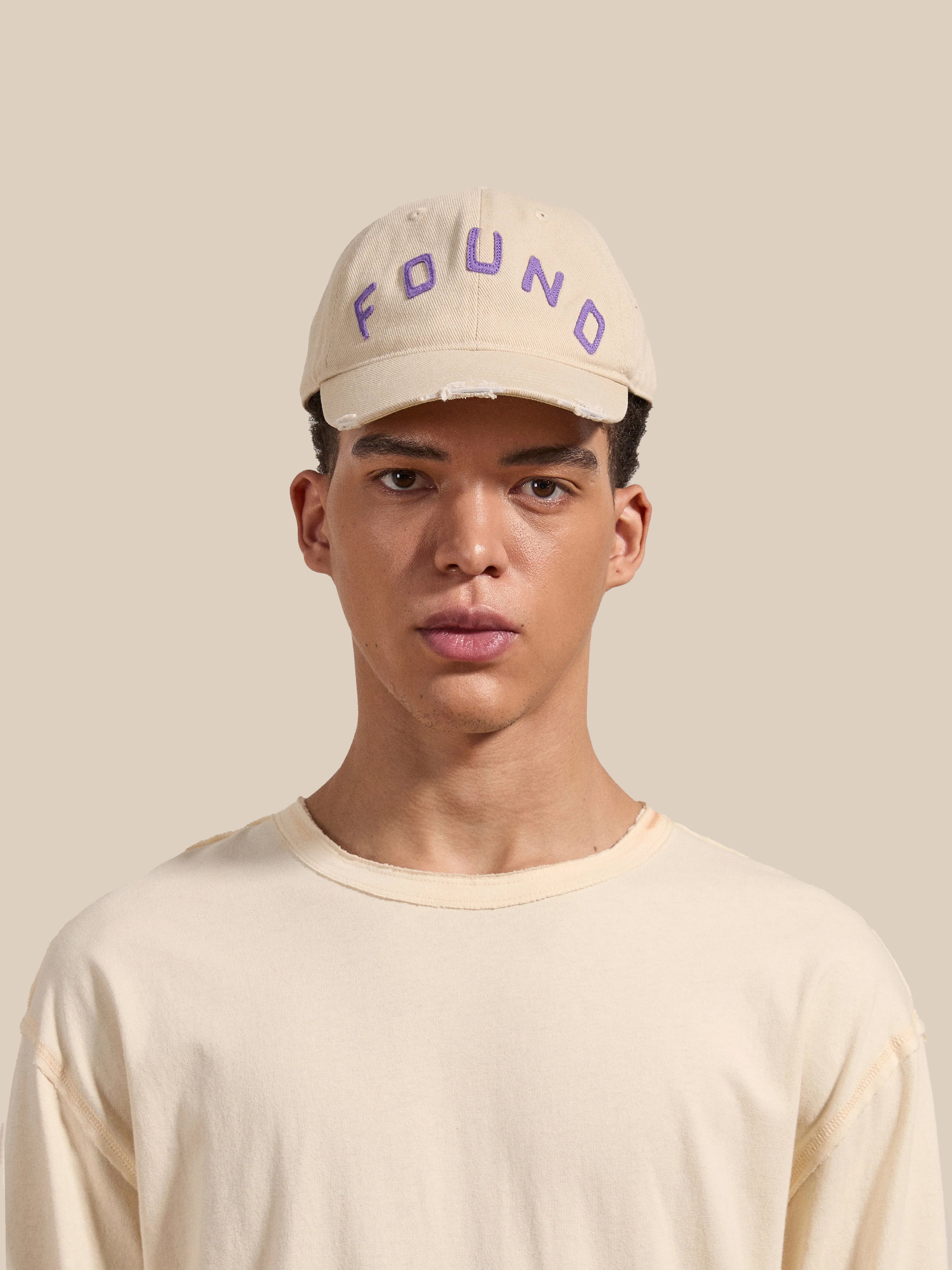 A young person dons a vintage washed fabric beige shirt and a stylish 6-panel curved brim Ivory Felt Patch Cap from the brand FOUND, set against a plain beige background.