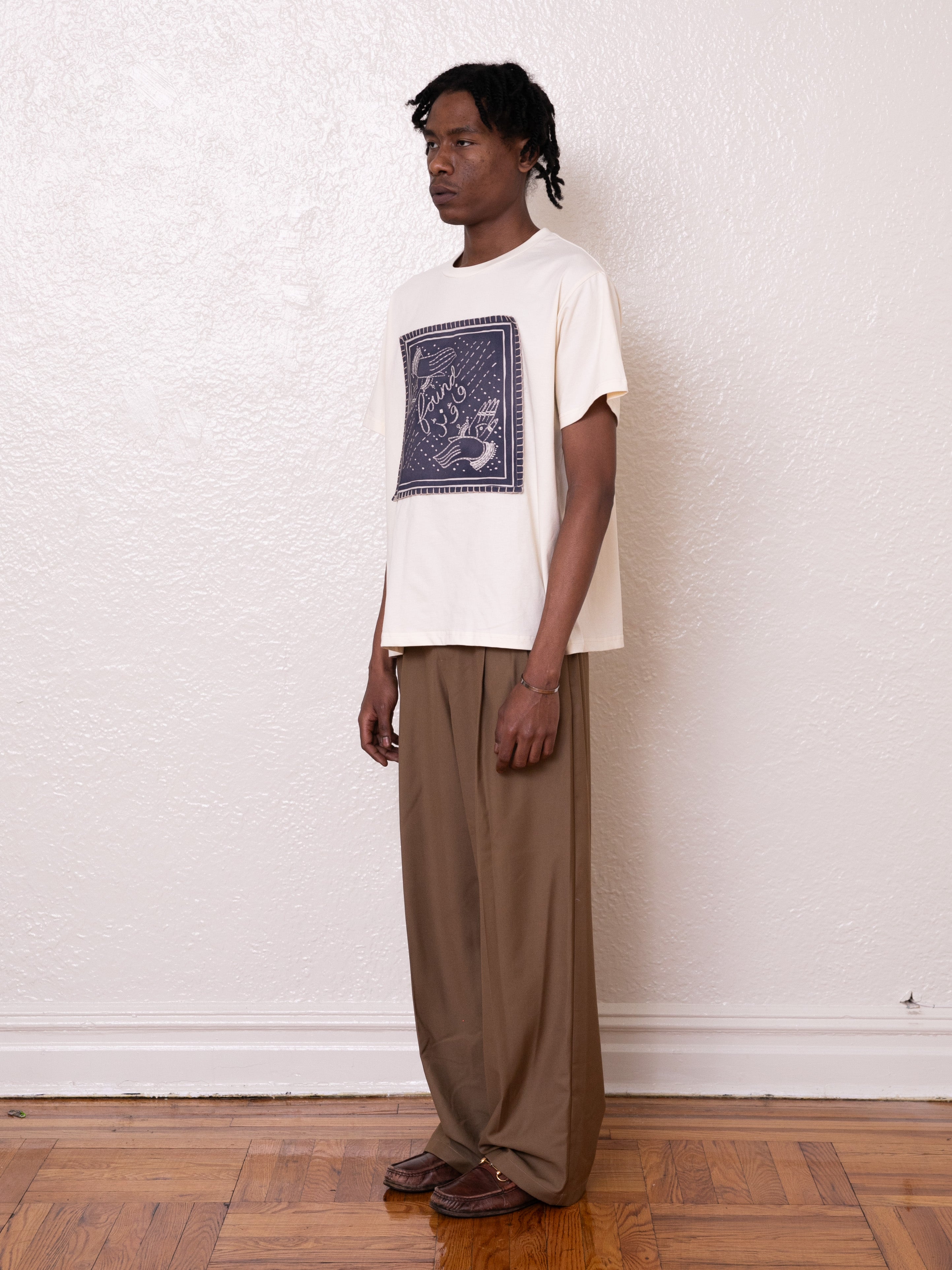 A person stands indoors against a plain wall, wearing a white Hand Art Graphic Tee by FOUND, brown pants, and brown shoes on a wooden floor.