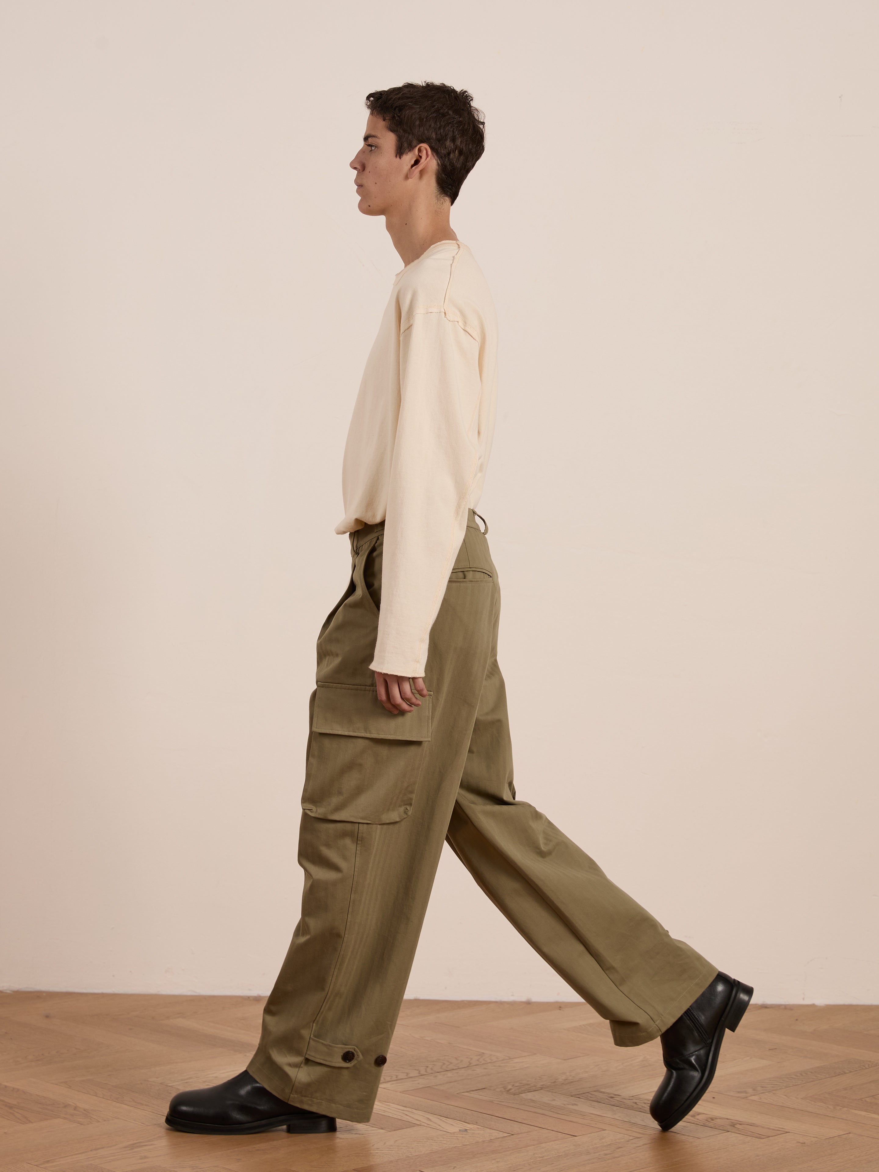 A person in a beige shirt and FOUND's Hava Herringbone Pleated Cargo Trousers walks right on a wooden floor against a plain background.