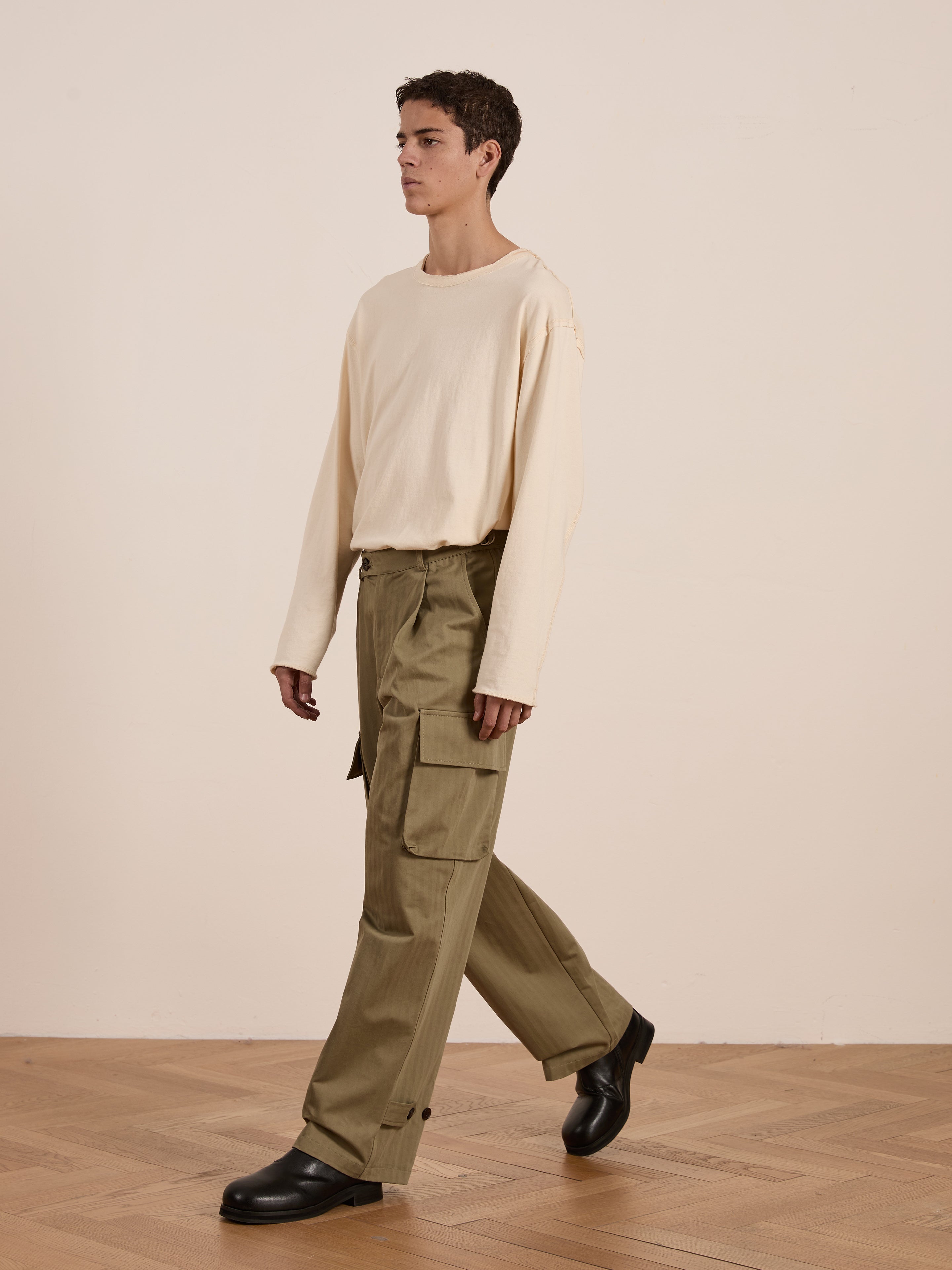 A person walks on a light wood floor wearing utility wear with a cream long-sleeve shirt and FOUND's Hava Herringbone Pleated Cargo Trousers in olive, paired with black shoes against a plain background.