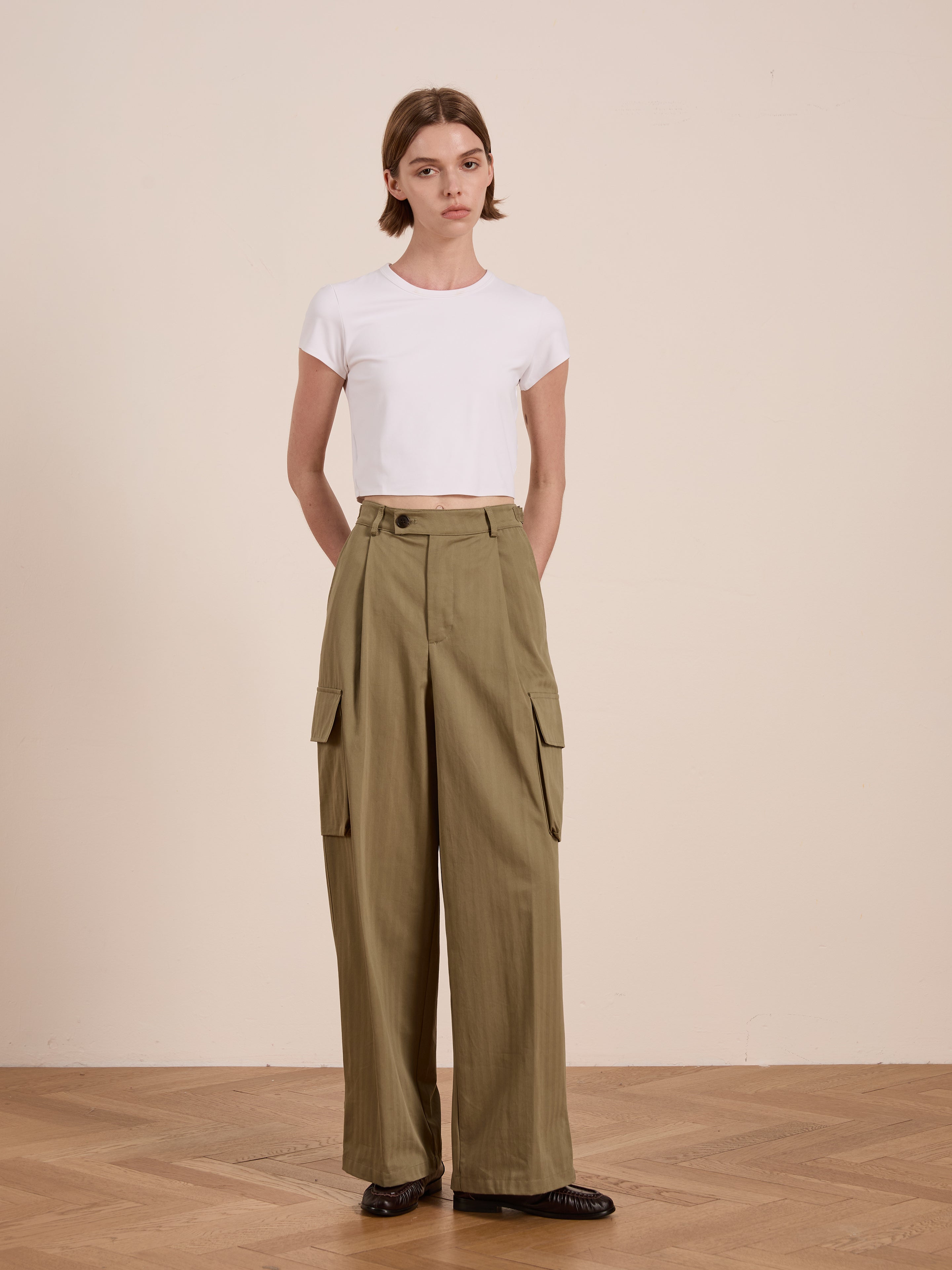 A person in a white crop top and FOUND's Hava Herringbone Pleated Cargo Trousers, crafted from cotton-blend fabric, stands against a neutral background.