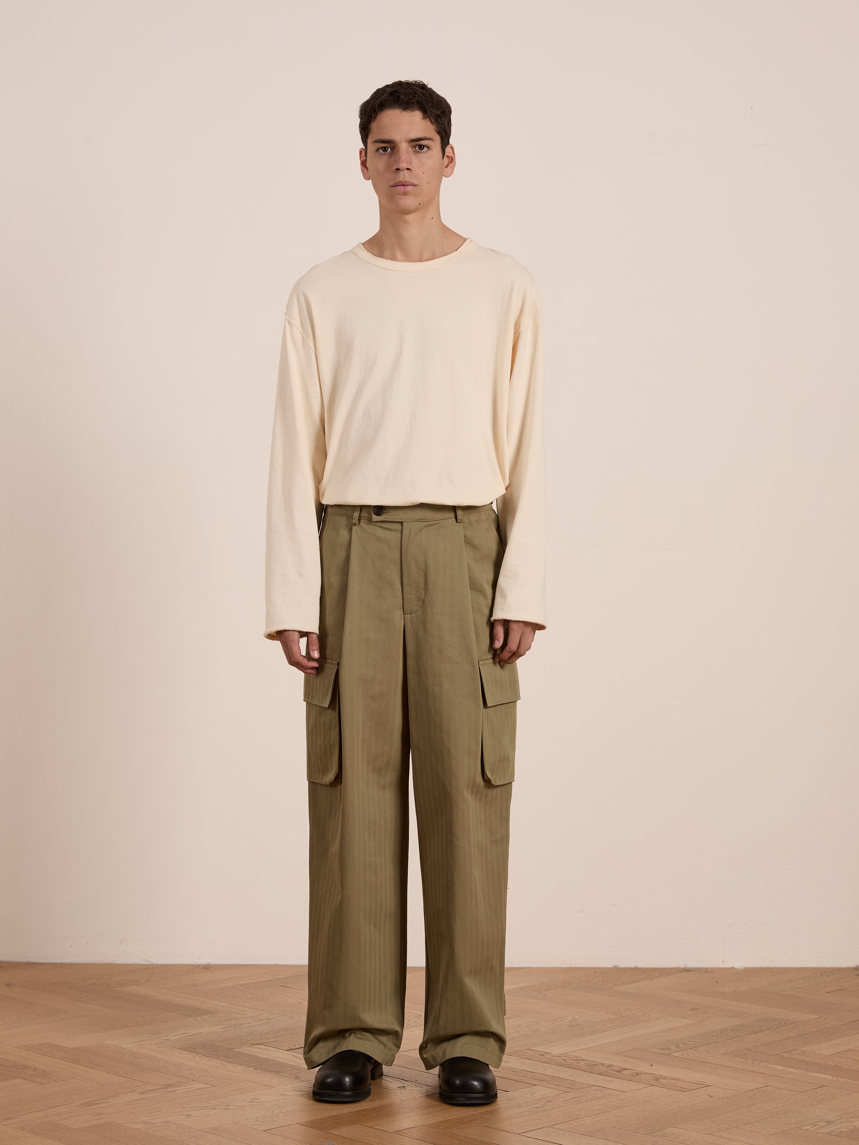 A person in a beige long-sleeve shirt, FOUND's Hava olive green herringbone pleated cargo trousers, and black shoes stands on a wooden floor against a plain light-colored wall.