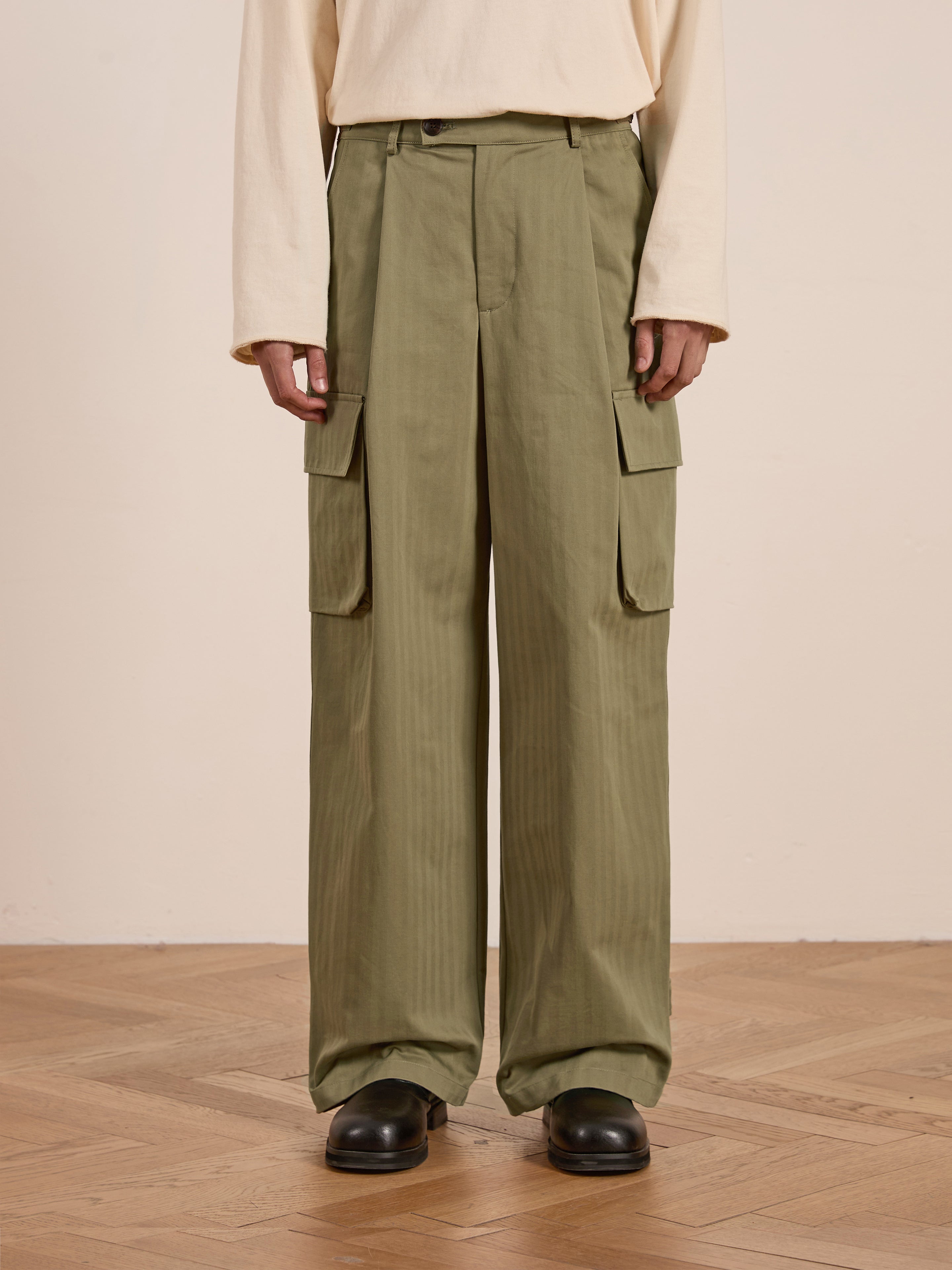 A person confidently stands on a wooden floor wearing FOUND's olive green Hava Herringbone Pleated Cargo Trousers and a beige long-sleeve shirt.