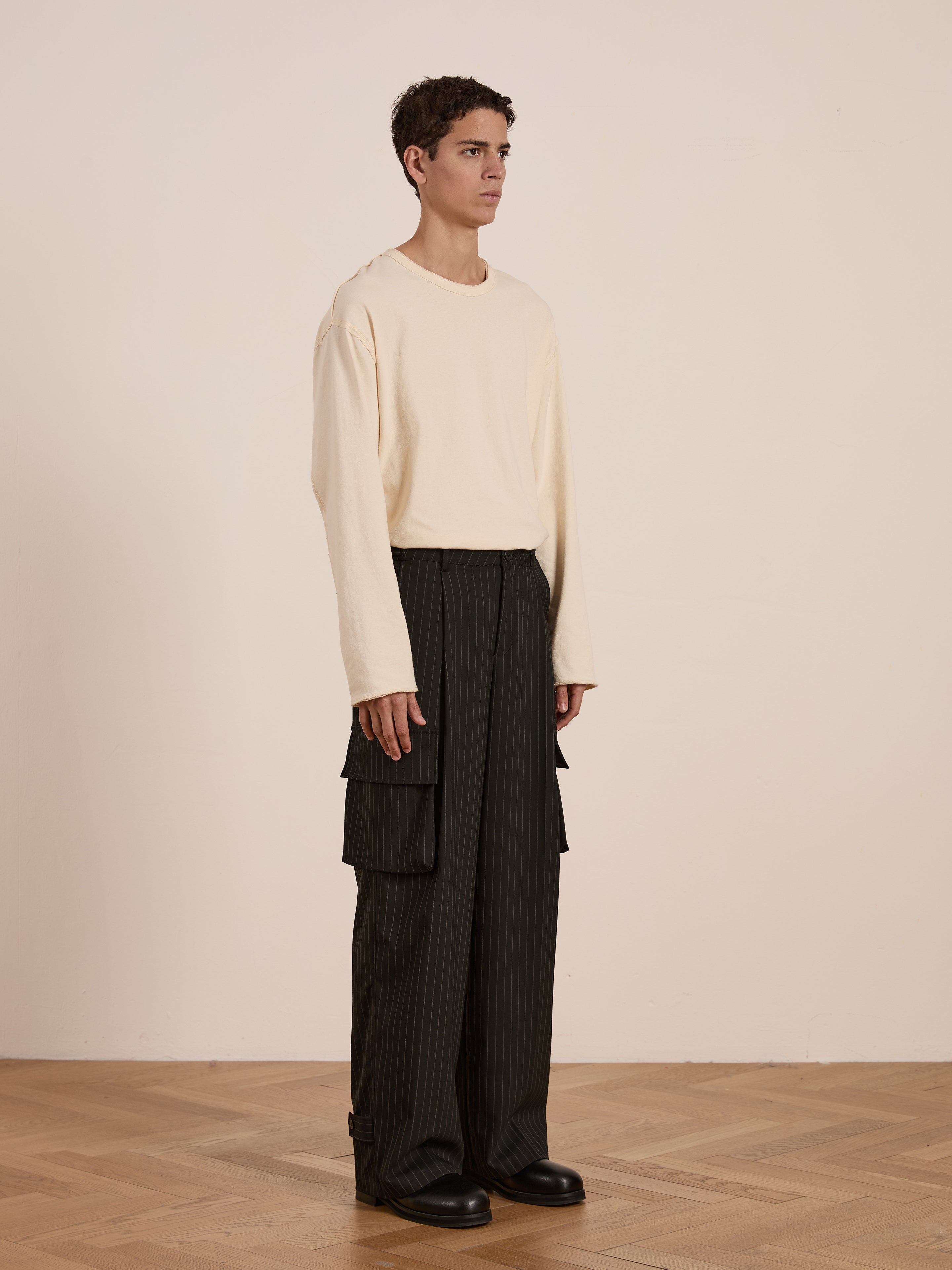 A person indoors wears a light long-sleeve shirt and the Hava Pinstripe Pleated Cargo Trousers by FOUND, made from wool-blend fabric with adjustable side tabs. They stand on a wooden floor against a beige wall.