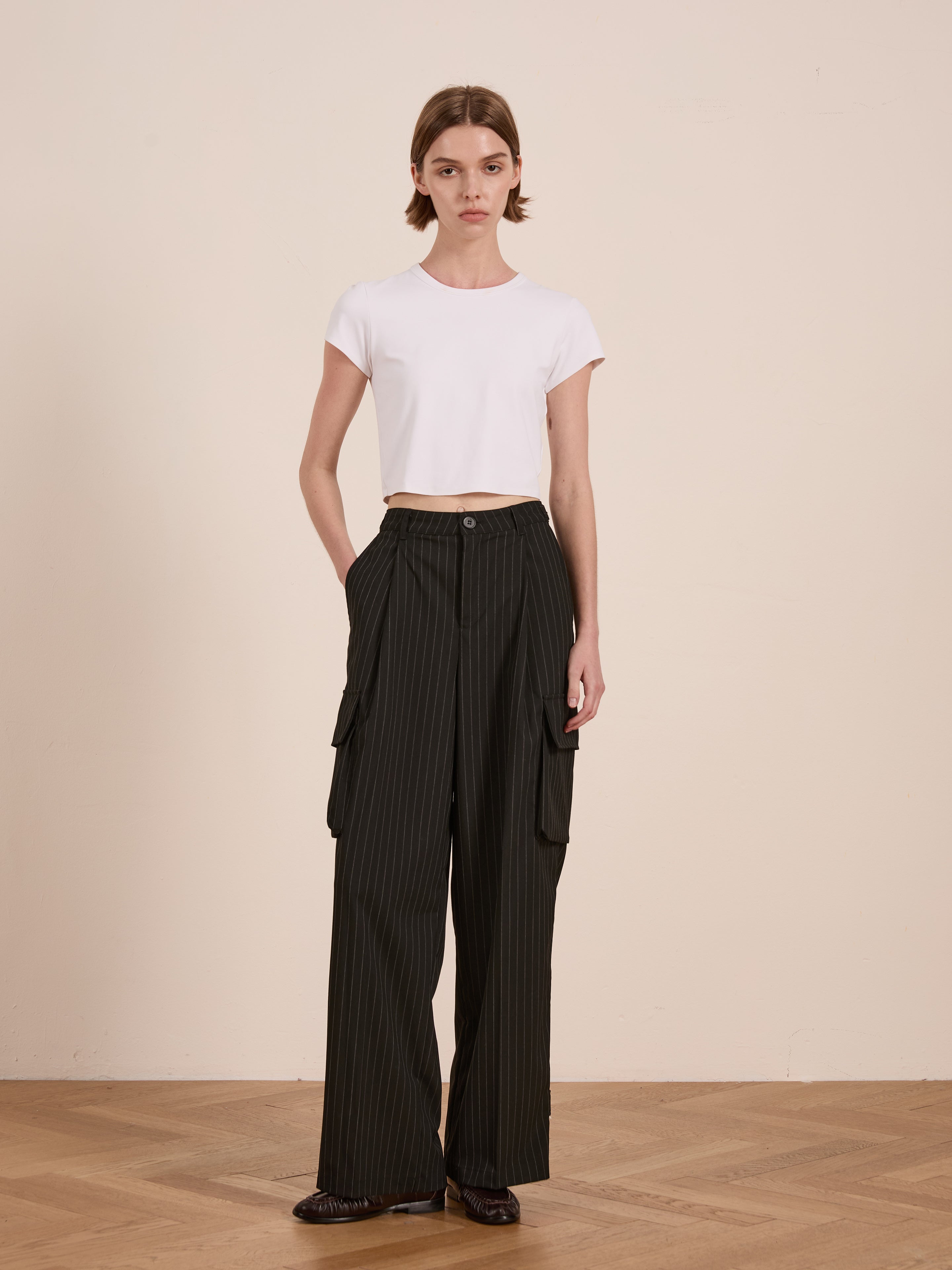 A person wears a white T-shirt with FOUND's Hava Pinstripe Pleated Cargo Trousers, showcasing sleek cargo detailing. The unisex relaxed fit pairs perfectly against a plain beige wall.