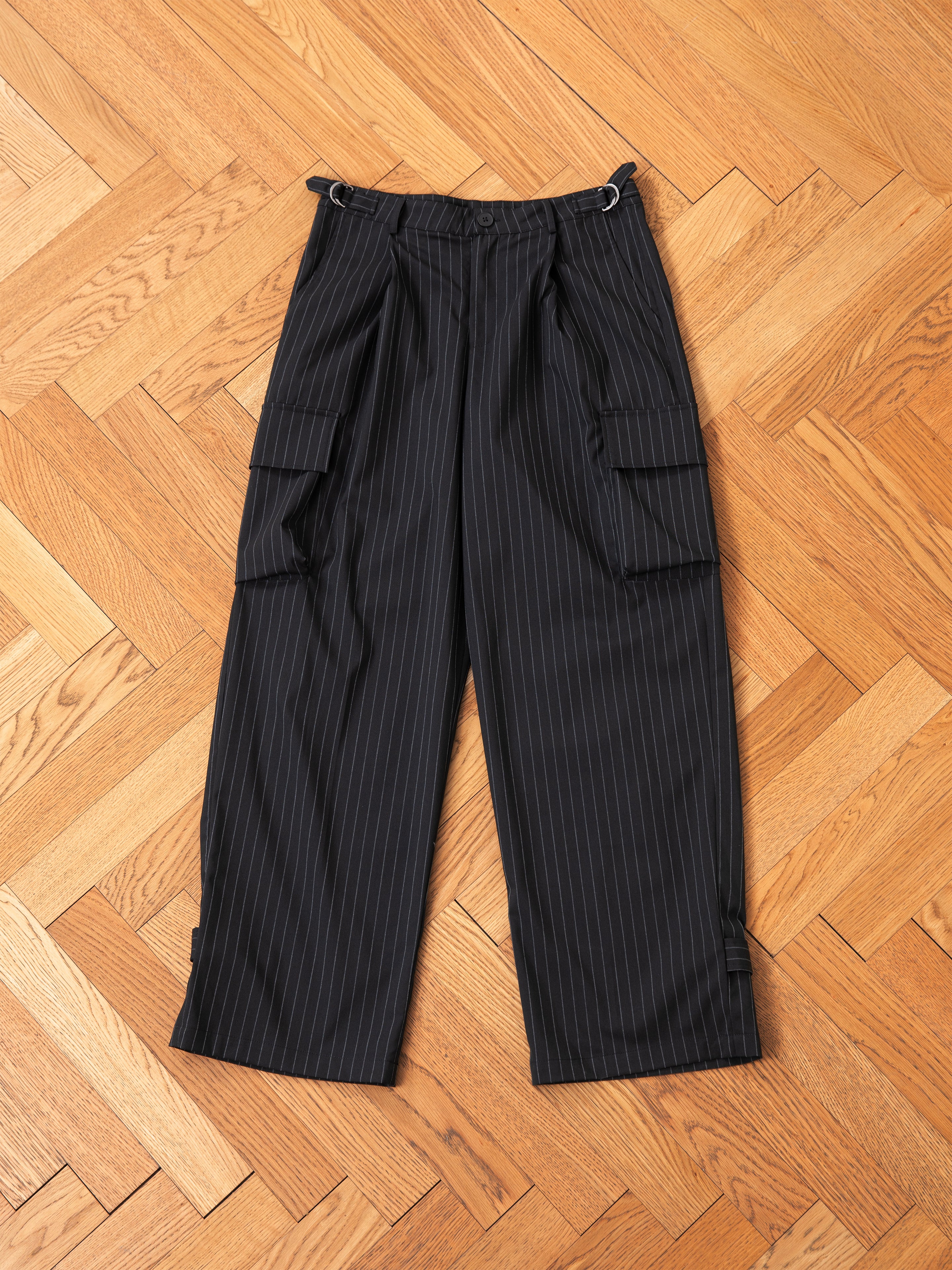 The Hava Pinstripe Pleated Cargo Trousers by FOUND, made from a black wool-blend with adjustable side tabs and multiple pockets, are laid flat on a wooden parquet floor.