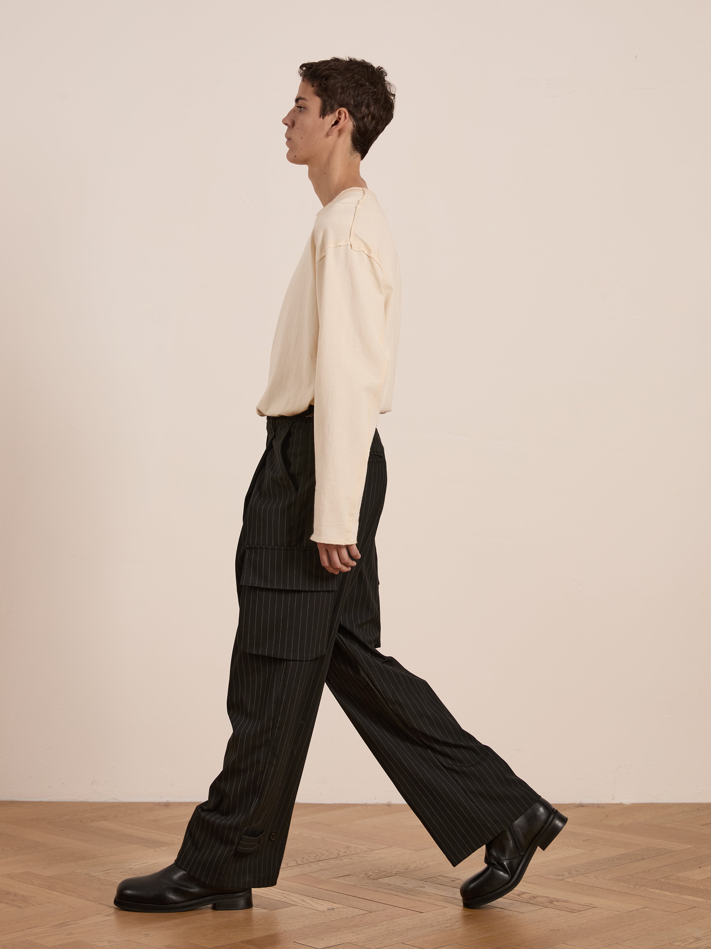 A person in a cream sweater and FOUND's Hava Pinstripe Pleated Cargo Trousers strolls on a wooden floor against a plain backdrop.